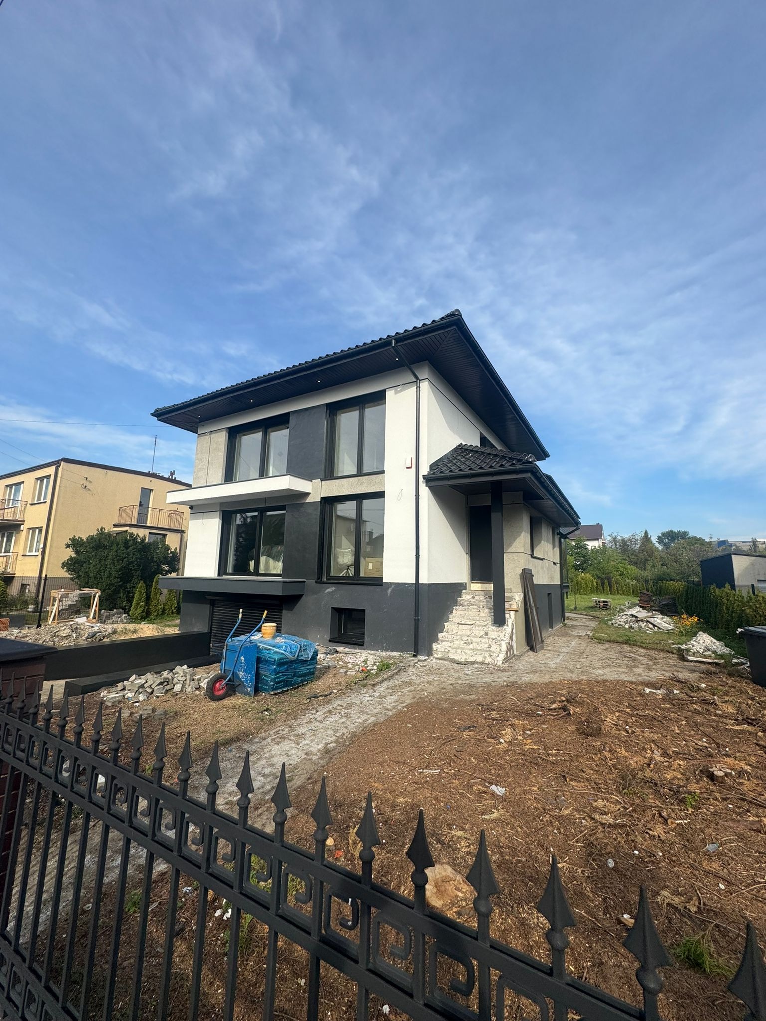 Dwukondygnacyjny dom z elewacją w odcieniach szarości i bieli, widoczny zza czarnego, ozdobnego ogrodzenia. Na pierwszym planie taczka z materiałami budowlanymi.