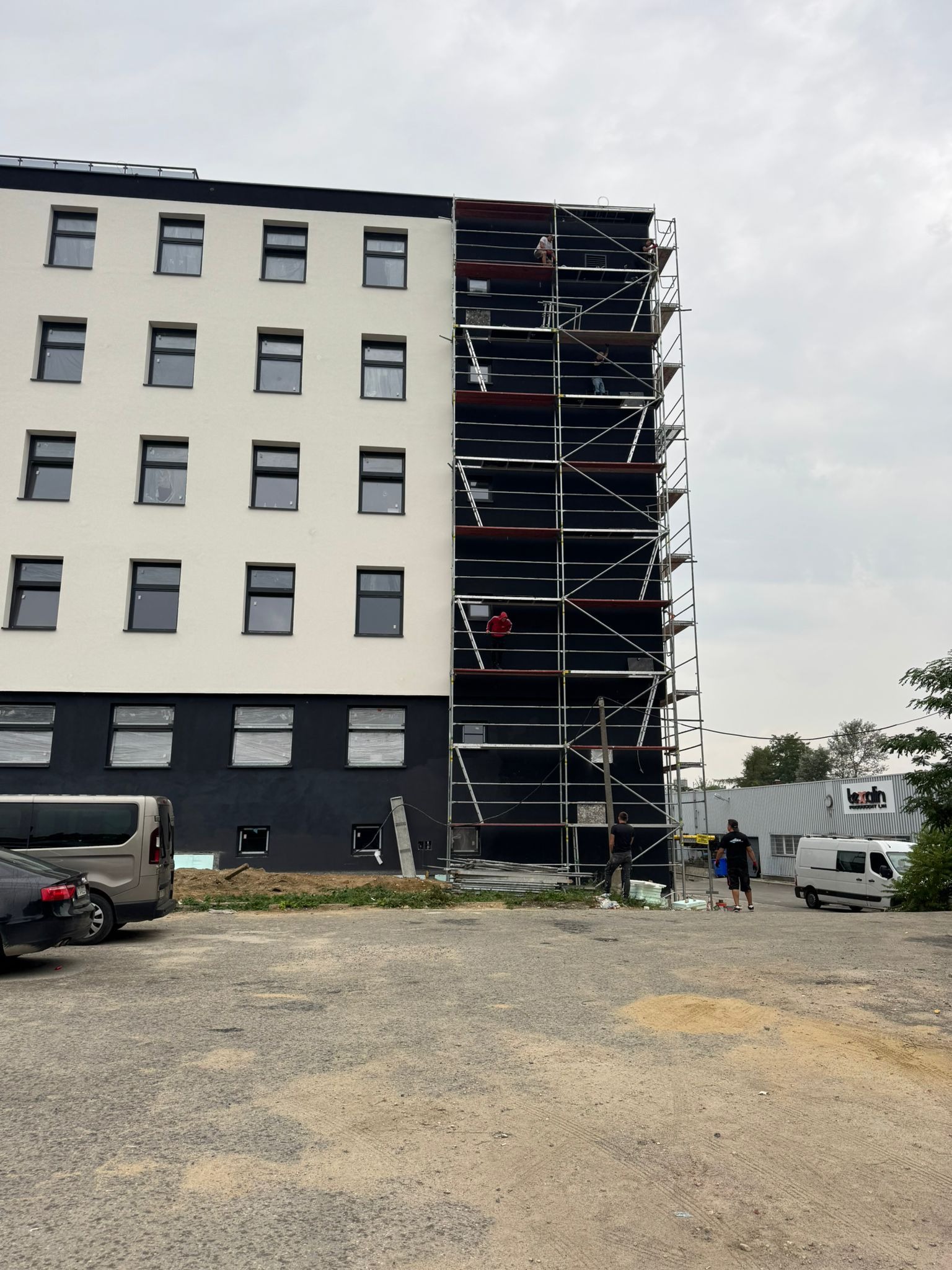 Budynek w trakcie prac elewacyjnych z rusztowaniem. Część fasady biała, część czarna. Widoczni pracownicy i zaparkowane samochody. Pochmurne niebo w tle.