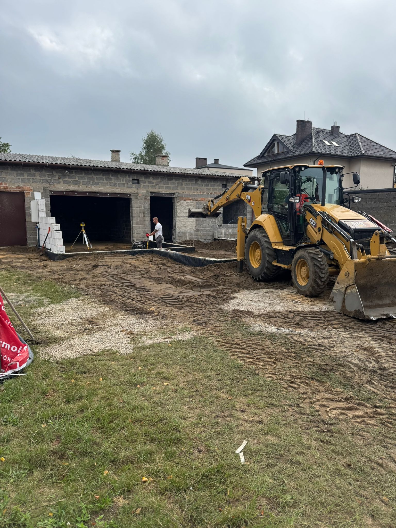 Żółta koparko-ładowarka CAT 432 w trakcie prac ziemnych przy budowie garażu z bloczków betonowych. Widoczny pracownik z niwelatorem laserowym.