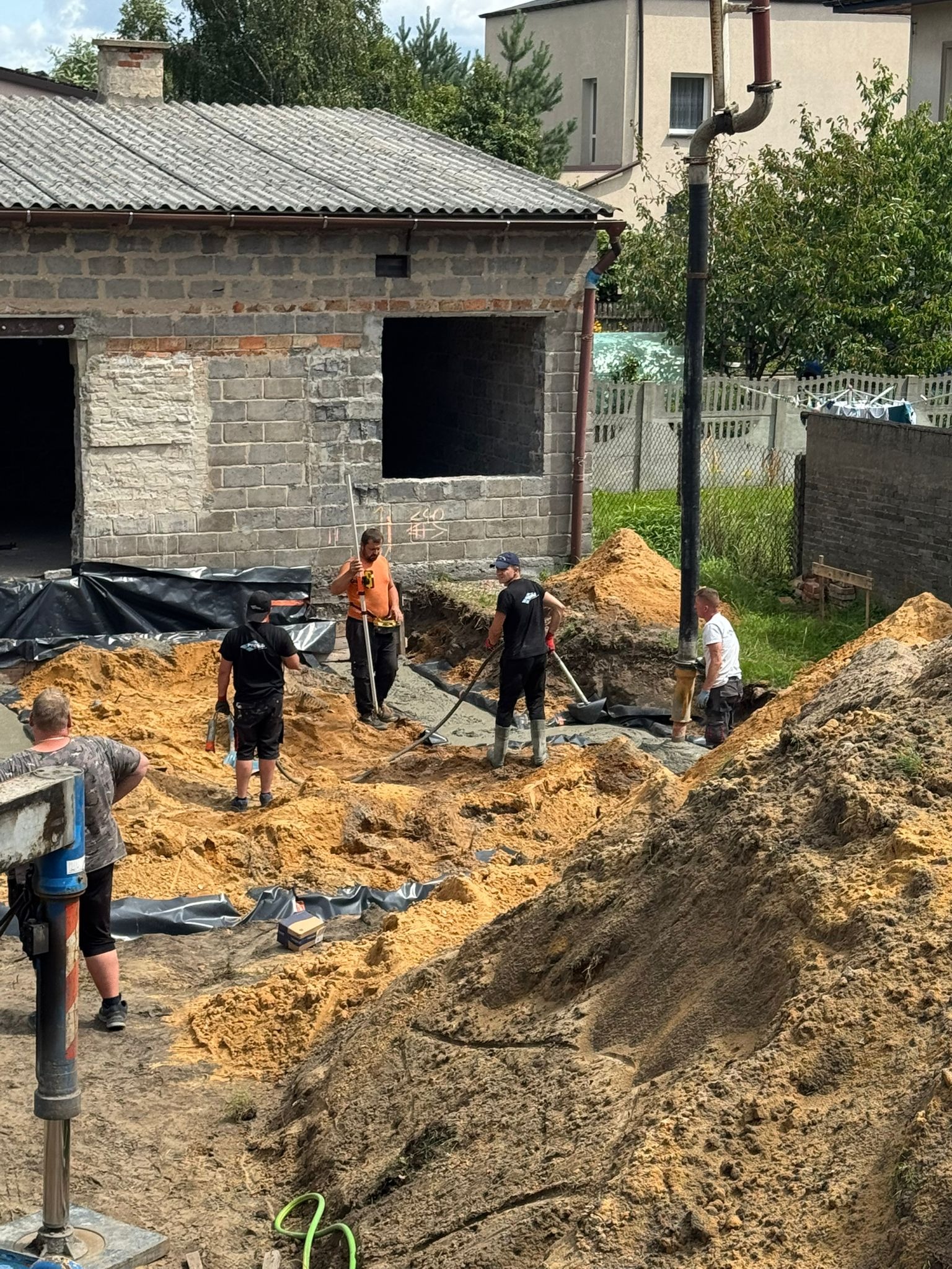 Prace ziemne przy fundamentach budynku z cegły. Widoczni robotnicy, sprzęt budowlany i wykop zabezpieczony folią. Dzień roboczy, naturalne oświetlenie.