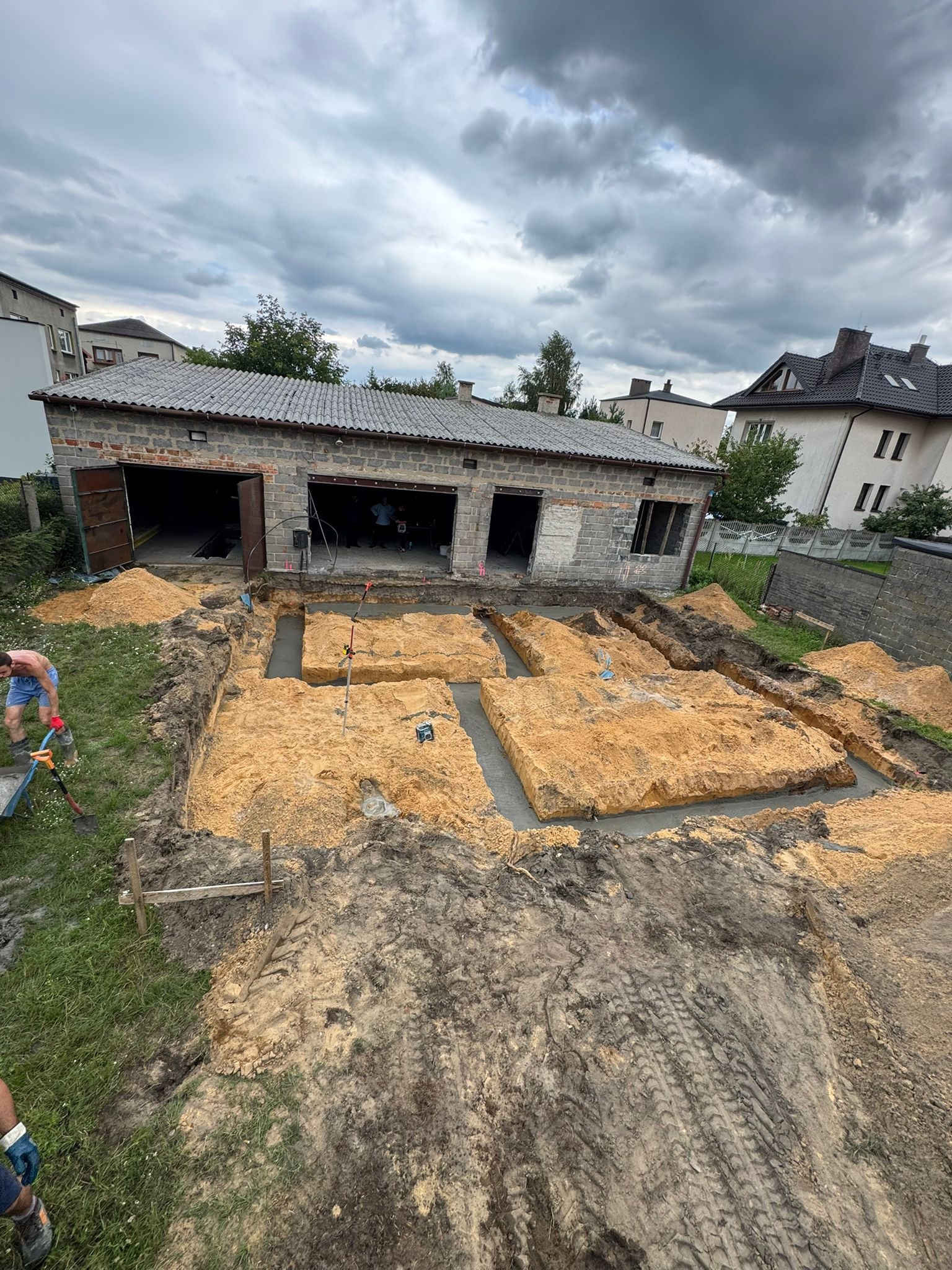 Wykopane fundamenty pod budynek z częściowo wylanym betonem, widoczne narzędzia i pracownik z taczką. Prace ziemne w toku, pochmurne niebo w tle.