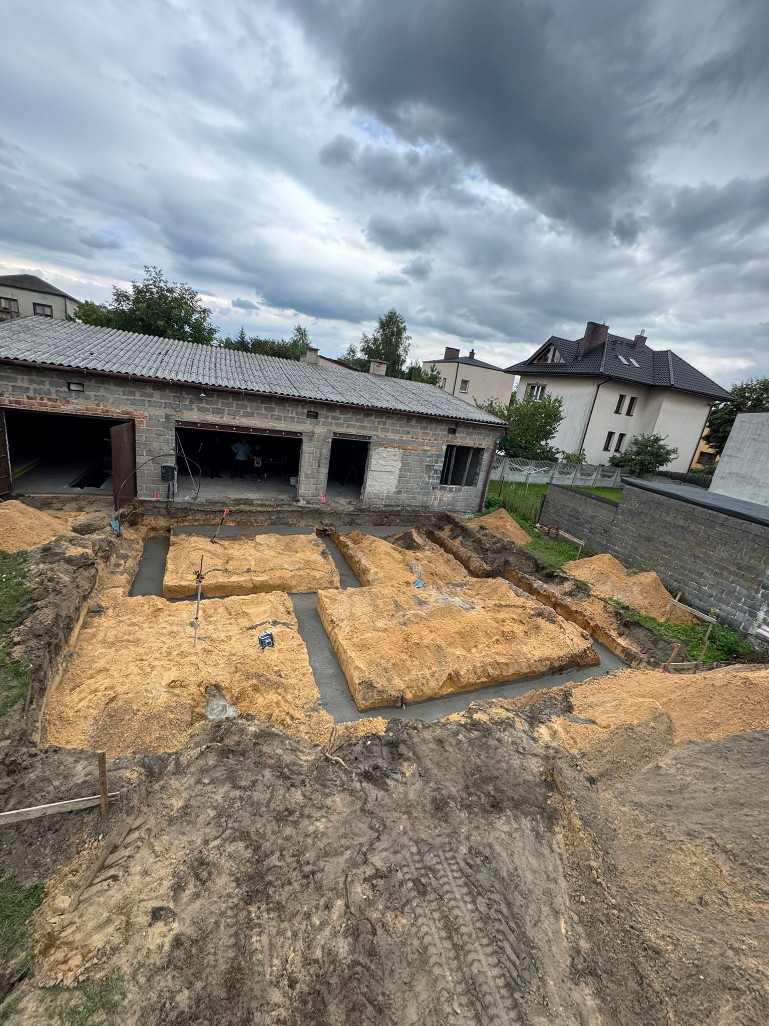 Wykopane fundamenty pod budynek z częściowo wylanym betonem, widok z góry na plac budowy w pochmurny dzień. W tle budynki mieszkalne i drzewa.
