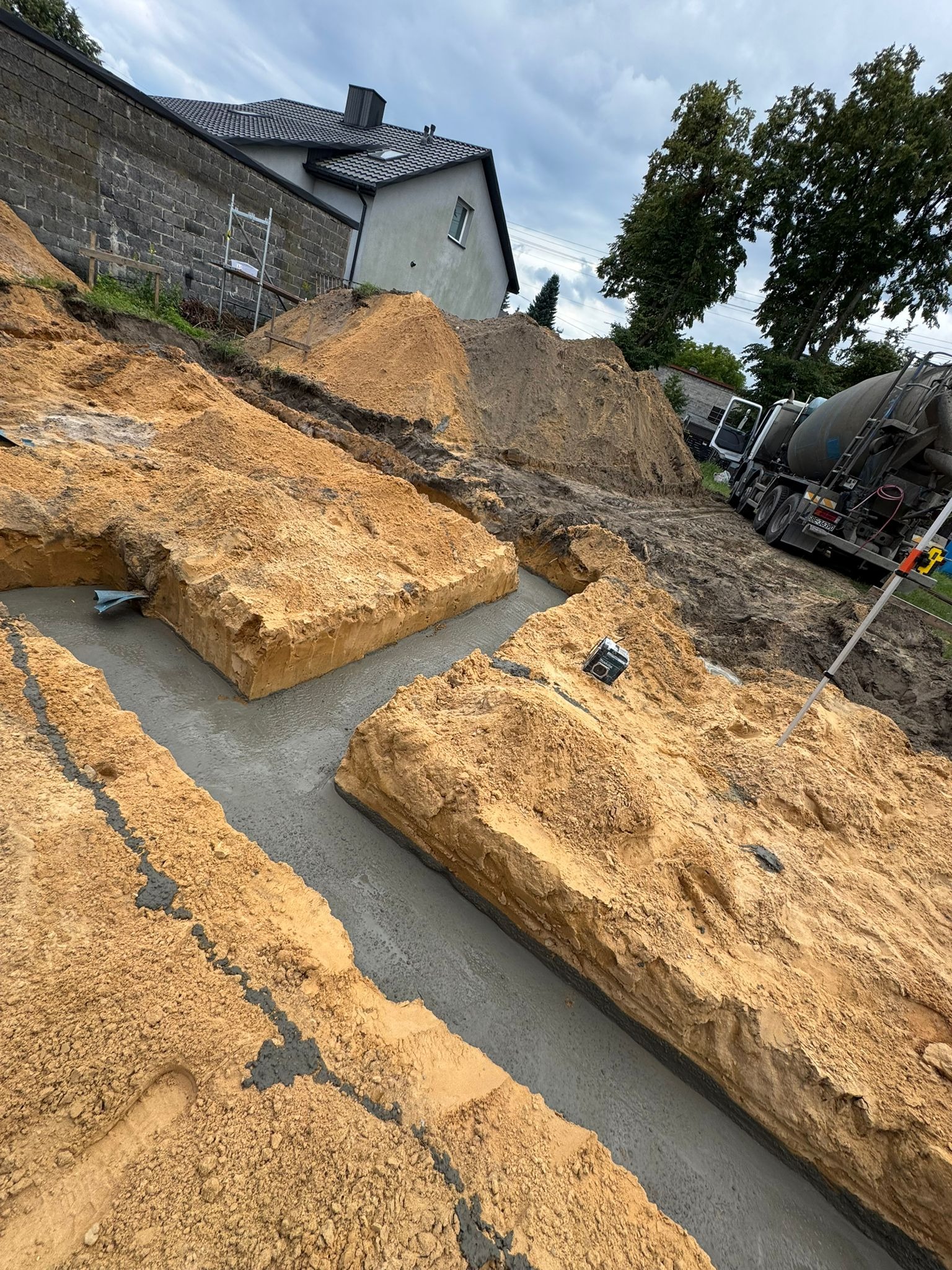 Wykopane fundamenty zalane betonem, widoczny mikser do betonu w tle. Prace ziemne i betonowe w trakcie realizacji. Ujęcie z góry, pochmurne niebo.