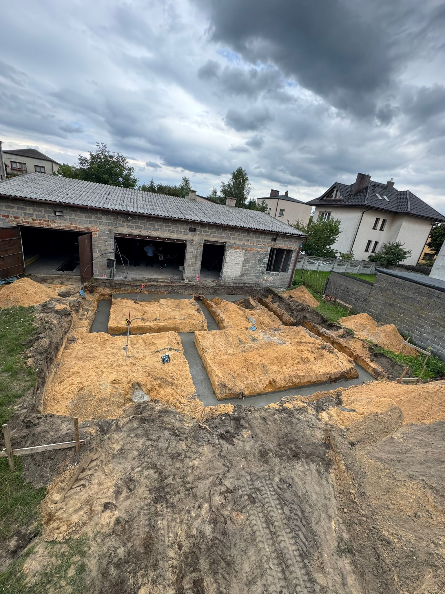 Wykopane fundamenty pod budynek z częściowo wylanym betonem, widoczne z góry. W tle budynek gospodarczy z otwartymi wrotami i pochmurne niebo.