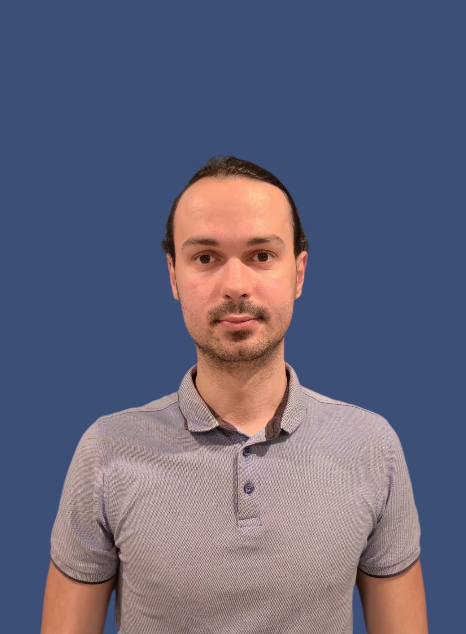 Portret mężczyzny w niebieskiej koszulce polo na jednolitym, ciemnoniebieskim tle. Ujęcie centralne, skupione na twarzy i górnej części tułowia. Neutralny wyraz twarzy.