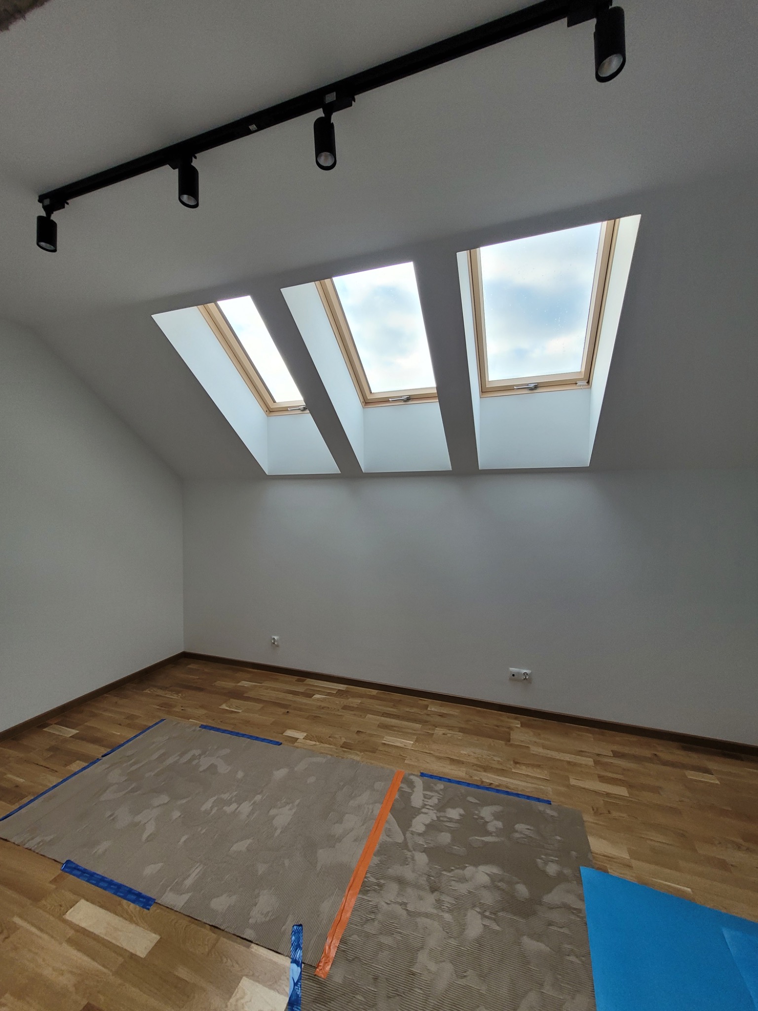 Pokój na poddaszu z trzema oknami dachowymi i parkietem. Podłoga zabezpieczona kartonem i folią malarską. Na suficie czarna listwa z reflektorami.