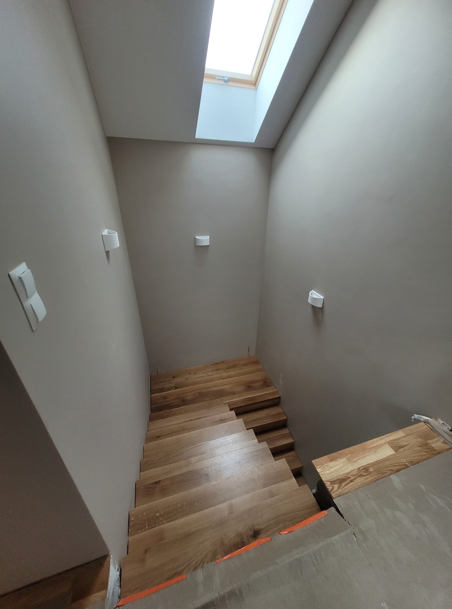 Jasne, drewniane schody w trakcie montażu, prowadzące w dół. Ściany w odcieniach szarości, oświetlenie punktowe. Widoczne elementy montażowe i okno dachowe.