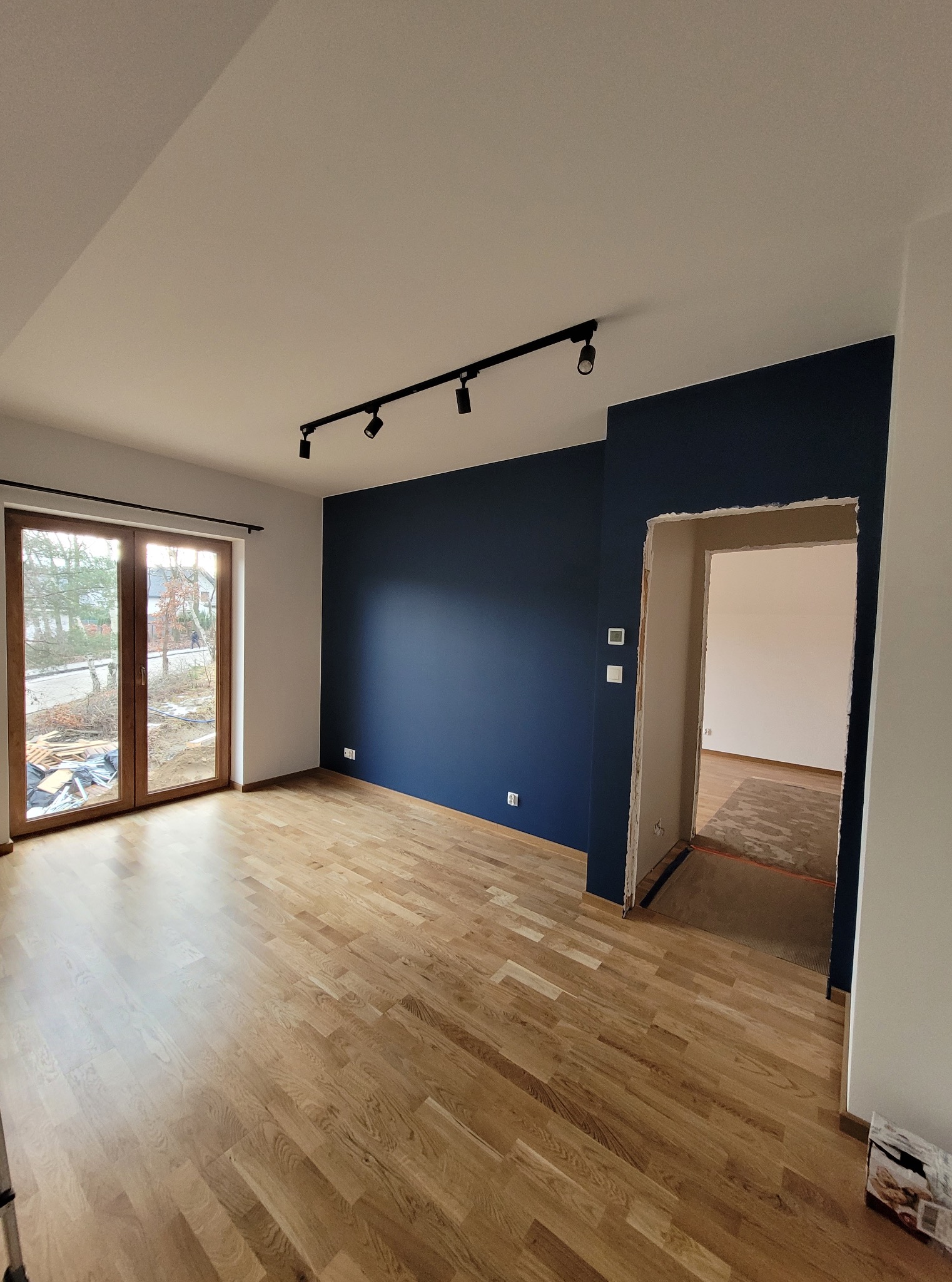 Jasny parkiet w przestronnym pokoju z granatową ścianą i nowoczesnym oświetleniem szynowym. Widoczne okno balkonowe i przejście do innego pomieszczenia.