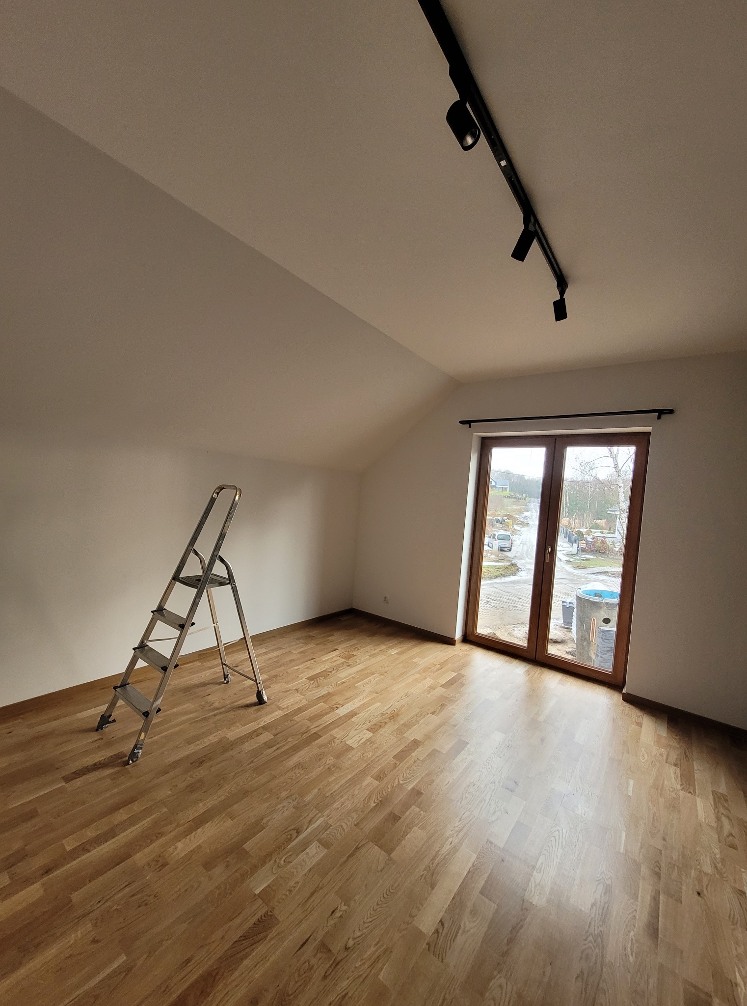 Jasne poddasze z drewnianą podłogą i drabiną malarską. Widoczne okno balkonowe i oświetlenie szynowe. Świeżo pomalowane ściany w neutralnym odcieniu.