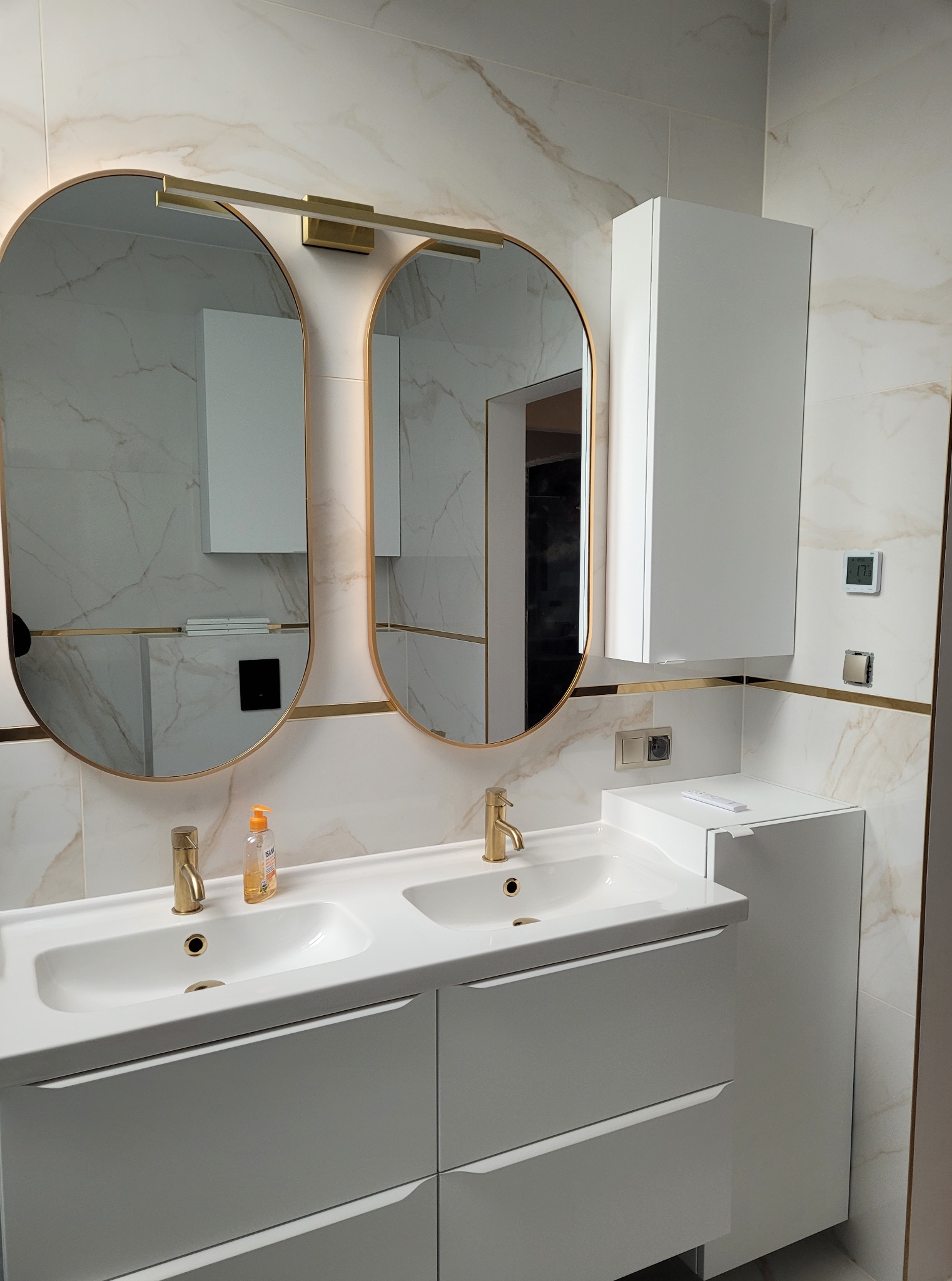 Wykończona łazienka z podwójną umywalką, złotą armaturą i owalnymi lustrami z podświetleniem. Białe szafki i marmurowe płytki tworzą elegancki wystrój.