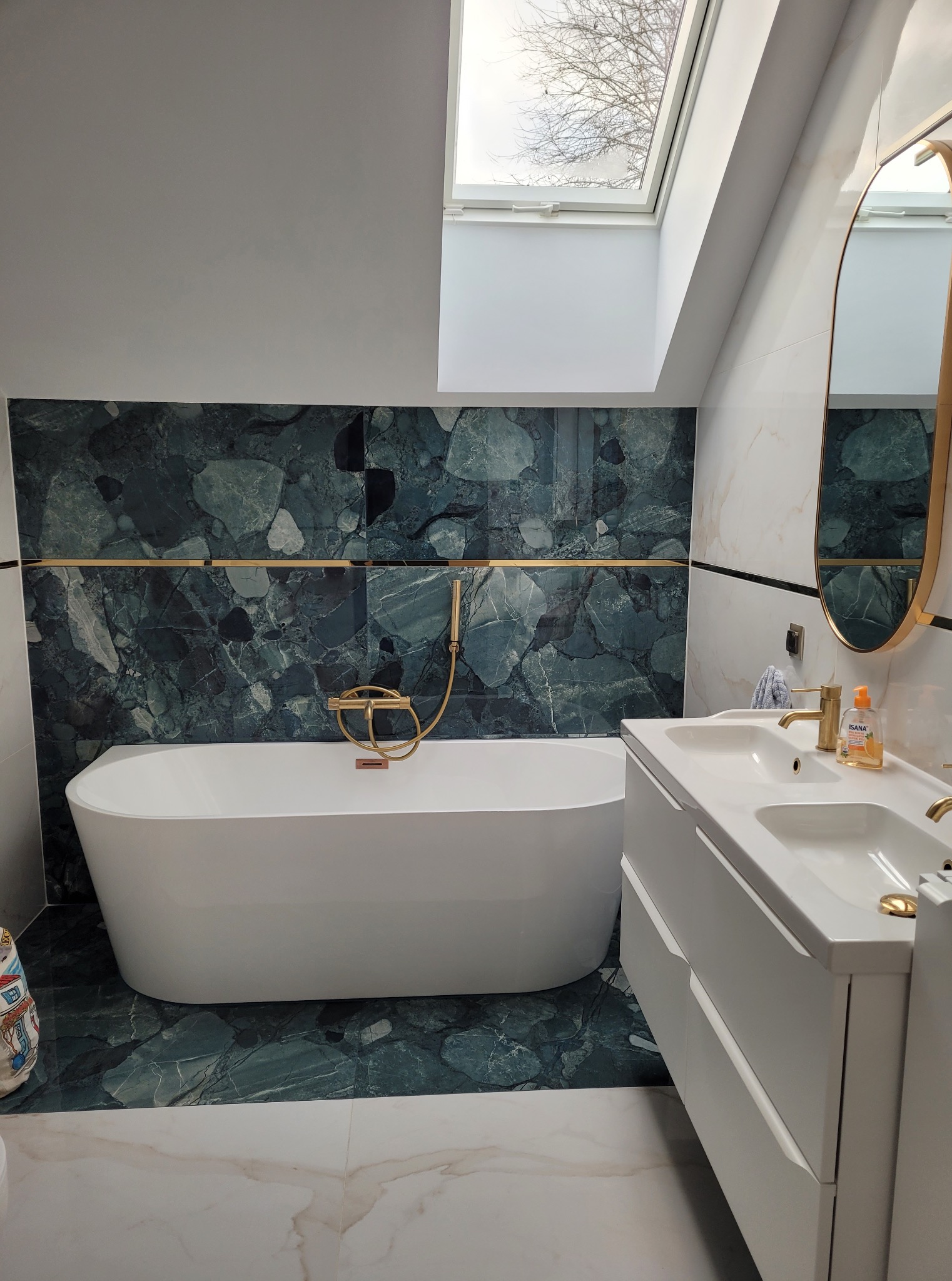 Nowoczesna łazienka z wanną wolnostojącą, zielonymi płytkami imitującymi kamień i złotą armaturą. Umywalka z szafką w bieli, lustro w złotej ramie i okno dachowe.
