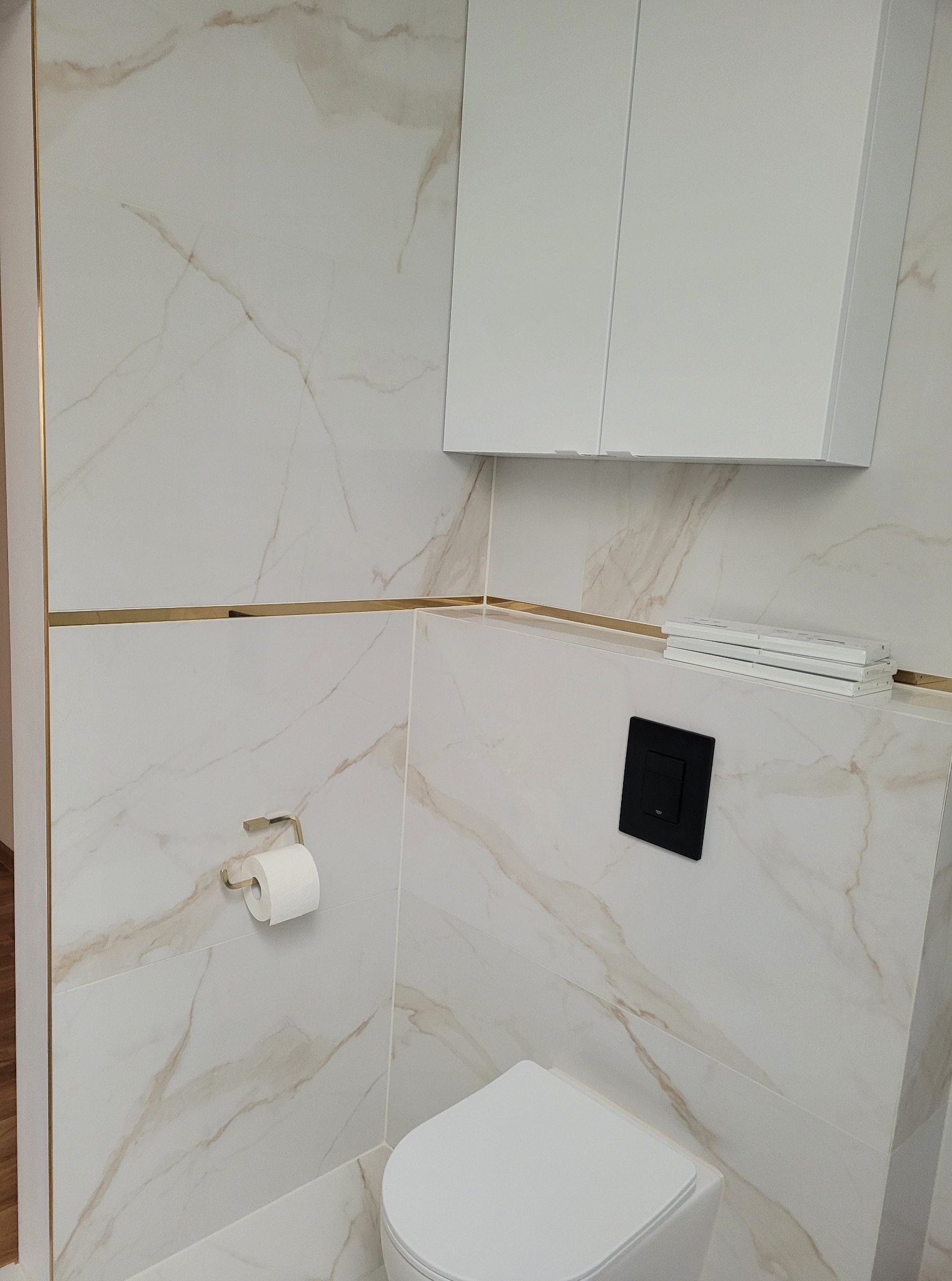 Elegancka toaleta z marmurowymi płytkami, złotymi akcentami i minimalistyczną zabudową. Nowoczesny design z czarnym przyciskiem spłukiwania i białym wyposażeniem.