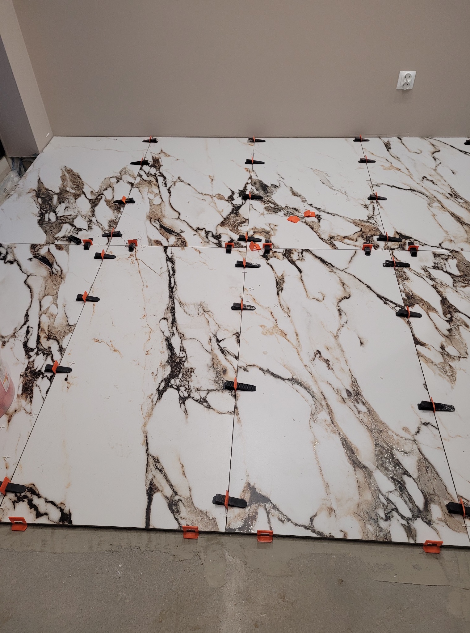 Układanie wielkoformatowych płytek imitujących marmur na podłodze, z użyciem klipsów poziomujących. Widoczny fragment sufitu i gniazdko elektryczne.