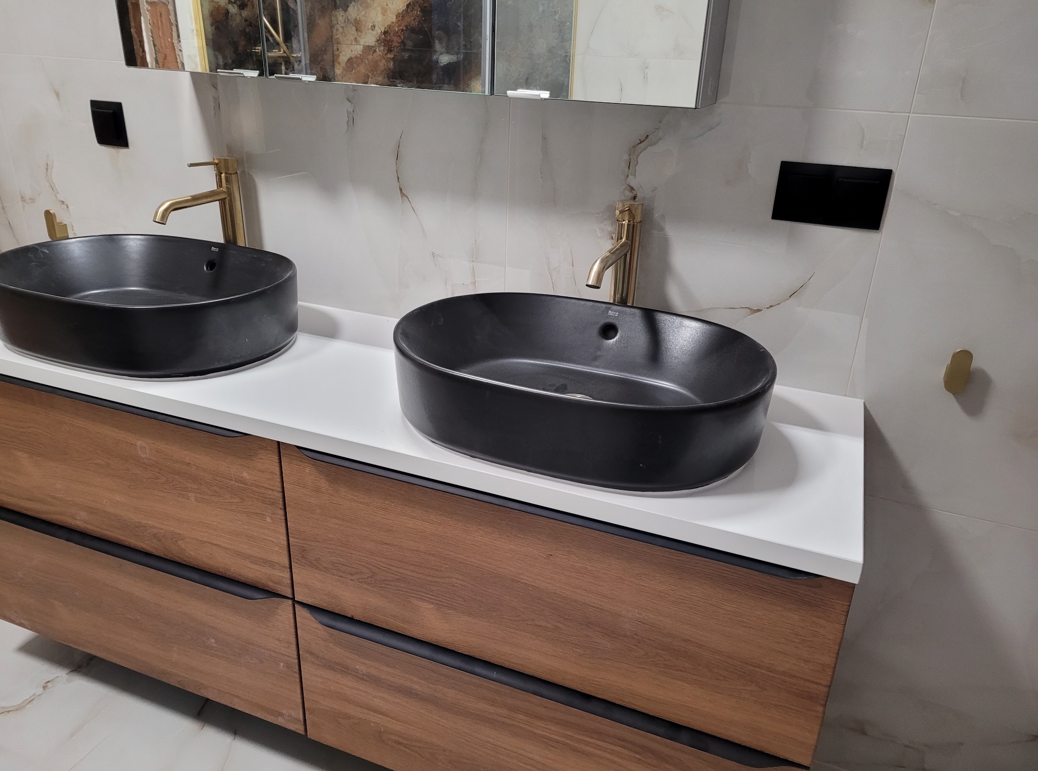 Nowoczesna łazienka z dwoma czarnymi umywalkami nablatowymi, złotymi bateriami i szafką z drewnianymi frontami. Marmurowe płytki na ścianie.