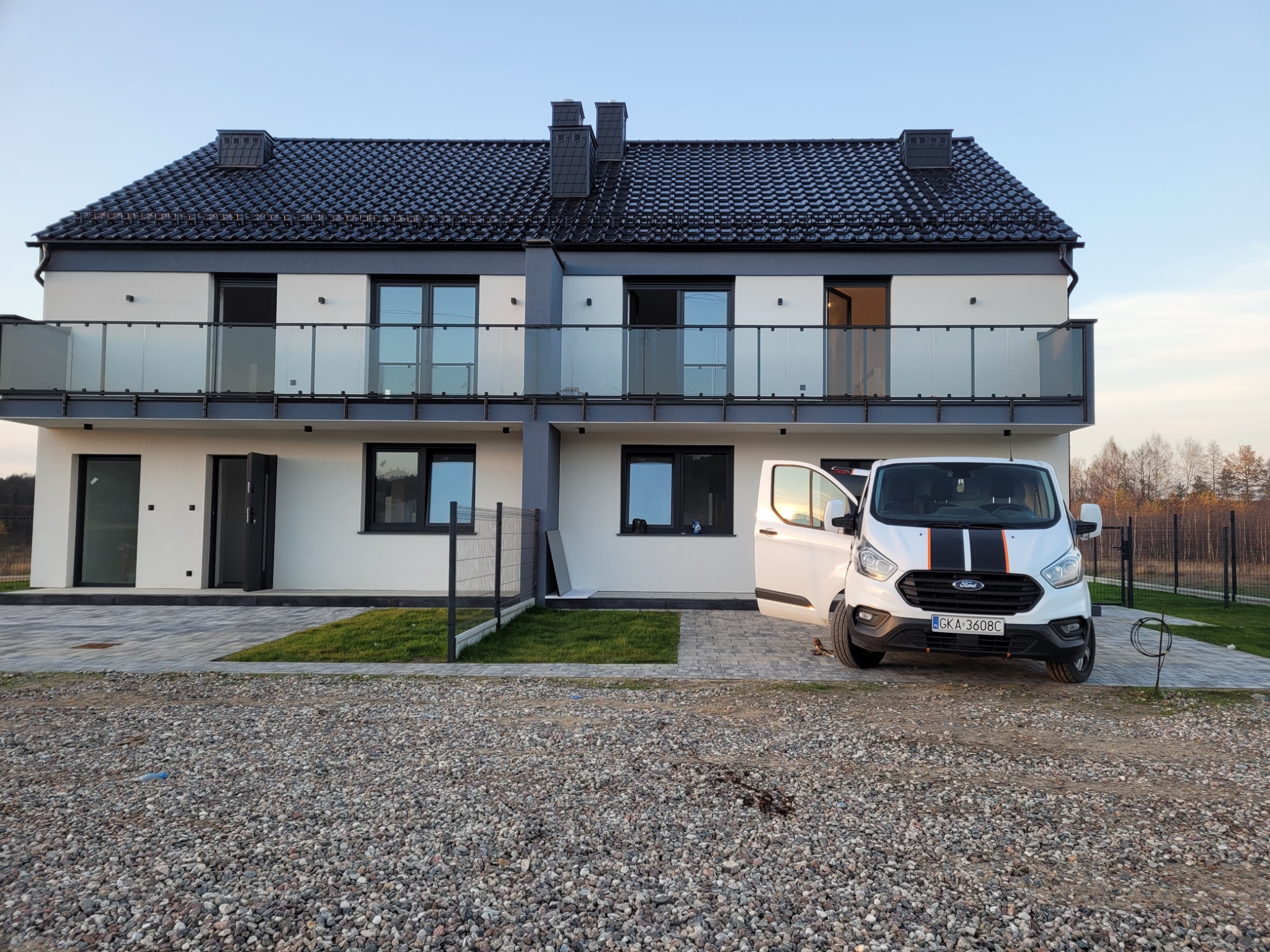 Dwukondygnacyjny dom bliźniak z balkonami i grafitową dachówką, na posesji stoi biały bus z otwartymi drzwiami. Widok z poziomu gruntu.