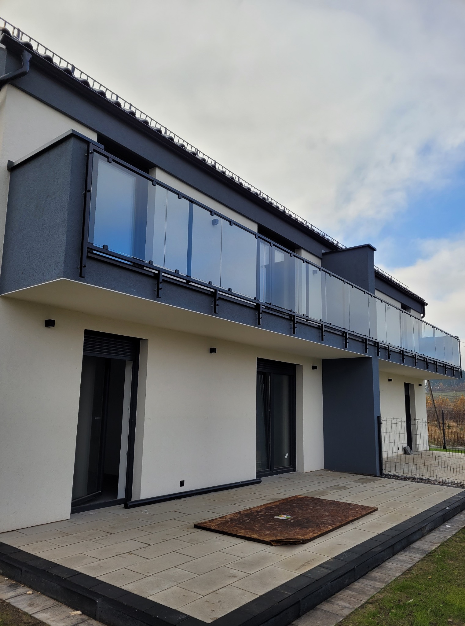 Nowoczesny budynek z balkonami z szklanymi balustradami i wyłożonym płytkami tarasem. Widok z dołu, podkreślający geometrię budynku i minimalistyczny design.