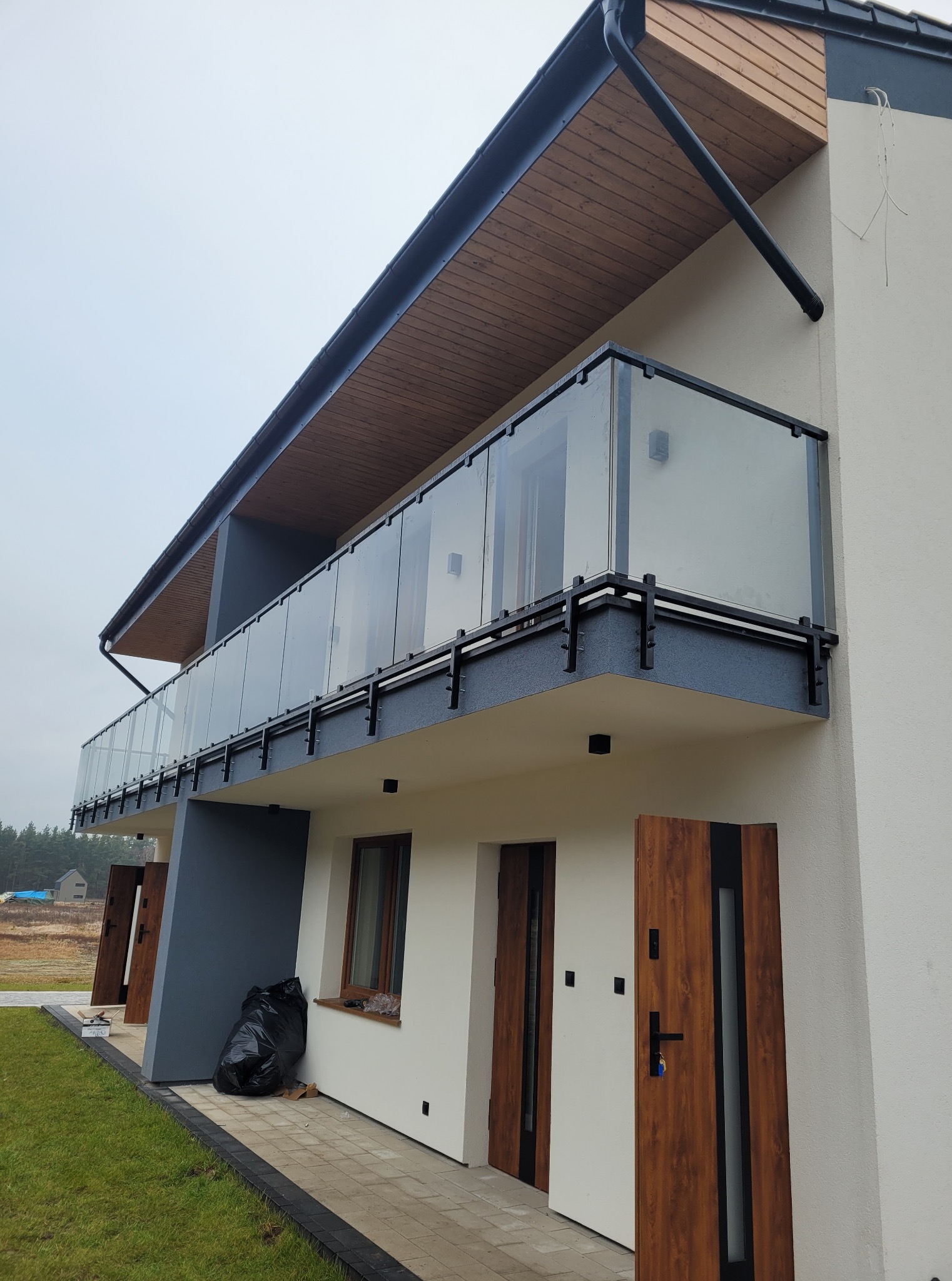 Nowoczesny budynek z balkonami z balustradami ze szkła i czarnymi ramami. Widoczne drewniane drzwi wejściowe i fragment trawnika. Minimalistyczny design.