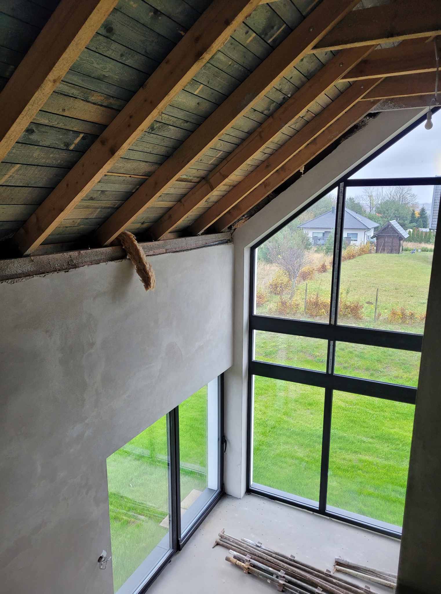 Surowy stan wnętrza z dużym oknem i widokiem na zewnątrz. Widoczne drewniane belki stropowe i fragment ocieplenia. Ściany w trakcie wykończenia, na podłodze leżą rury.