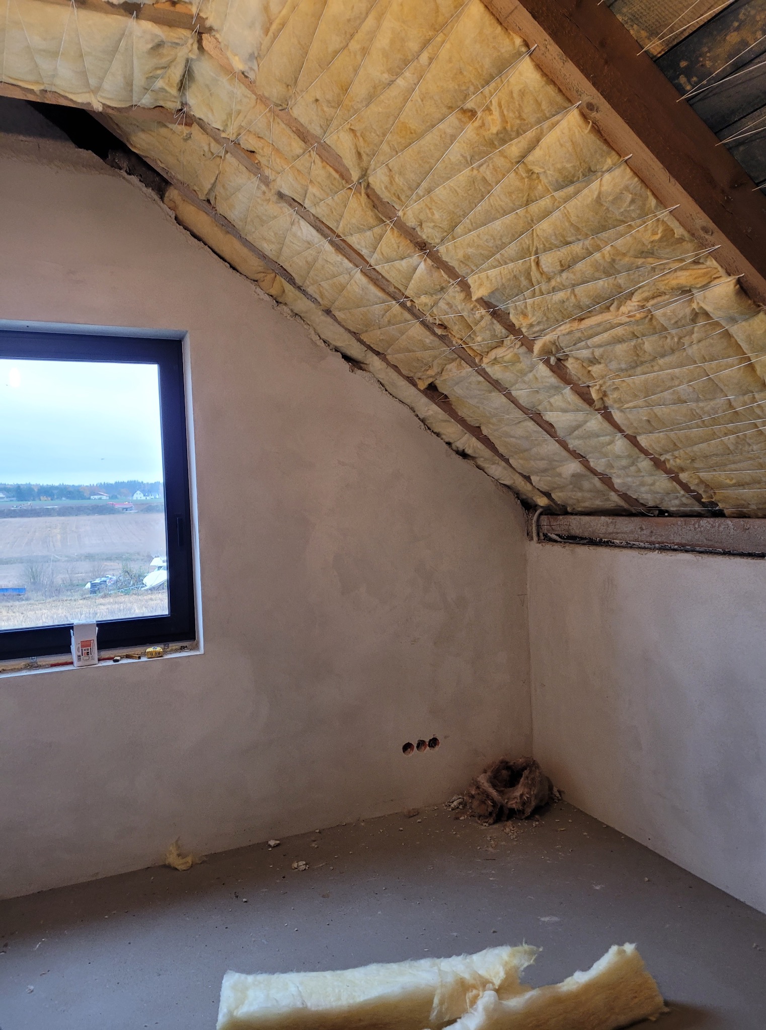 Poddasze w trakcie ocieplania wełną mineralną, widoczne drewniane belki stropowe i okno z widokiem na pole. Ściany przygotowane do dalszych prac wykończeniowych.