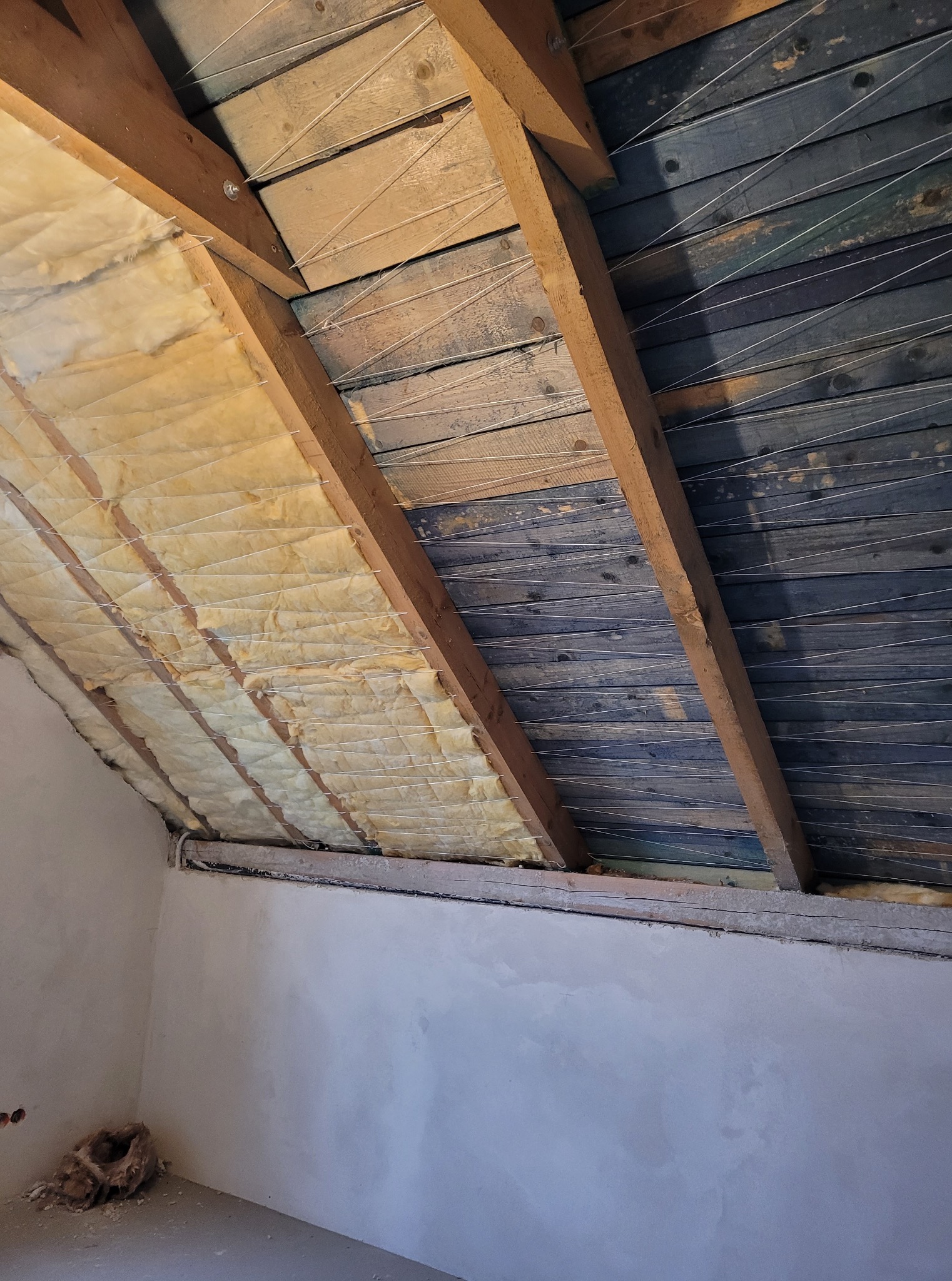 Ocieplenie poddasza wełną mineralną, widoczne drewniane belki konstrukcyjne i sznurki mocujące izolację. Ściany w stanie surowym, przygotowane do dalszych prac.