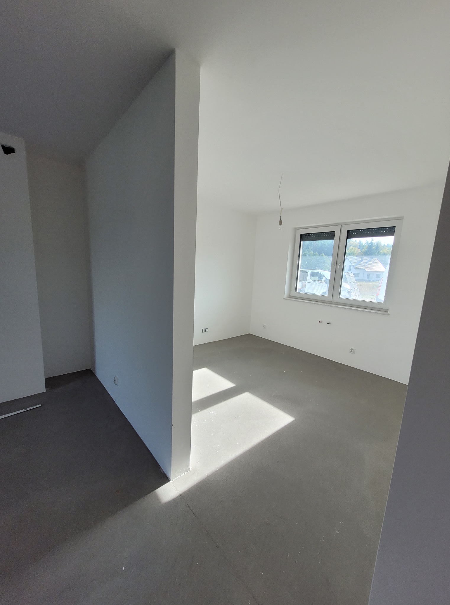 Jasne, puste wnętrze po wylewce, z białymi ścianami i oknem z widokiem na zieleń. Promienie słoneczne podkreślają gładką powierzchnię podłogi. Minimalistyczny design.