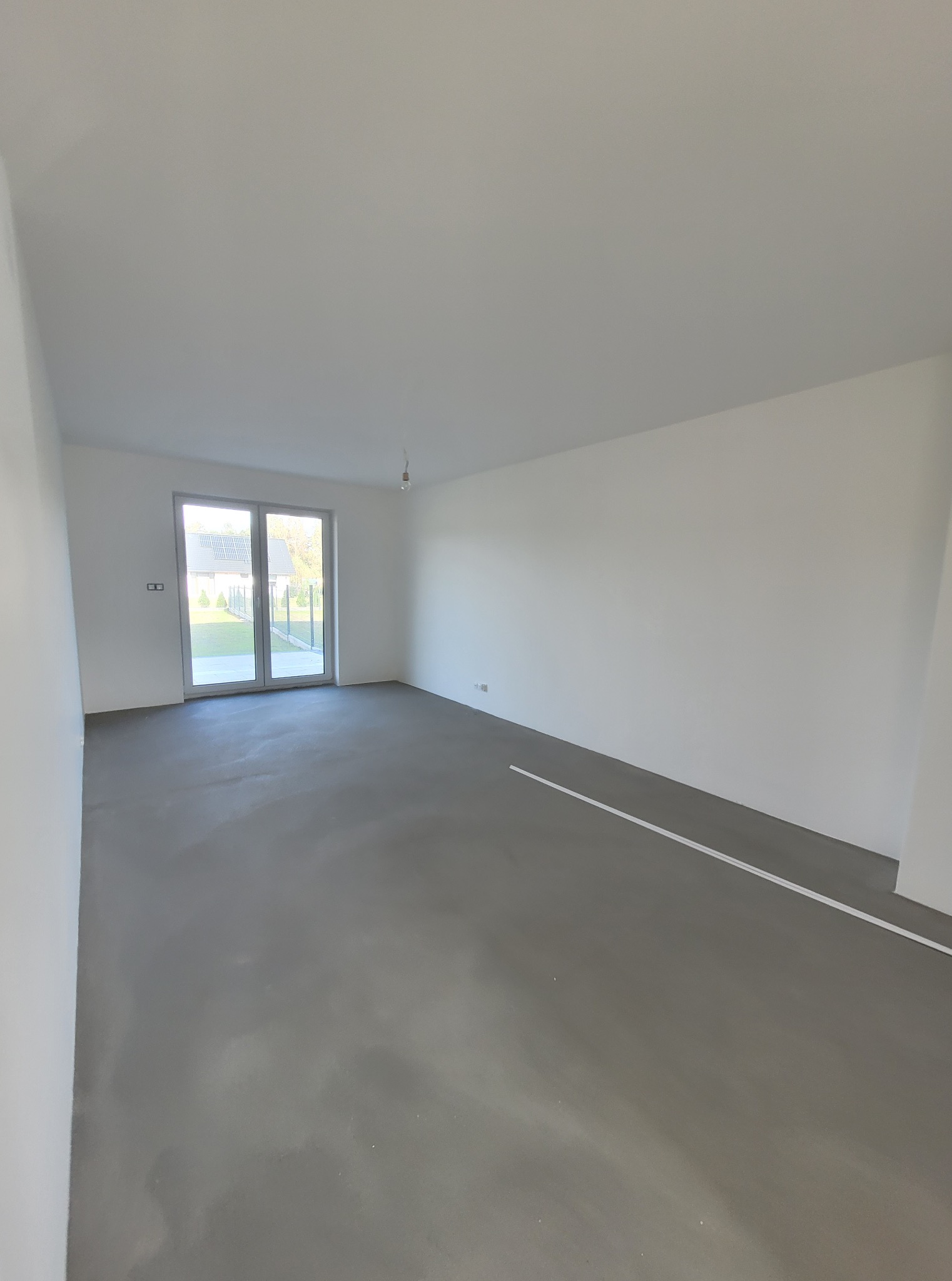 Wnętrze pokoju po wylewce, z widocznymi białymi ścianami i dużym oknem balkonowym. Podłoga szara, surowa, przygotowana do dalszych prac wykończeniowych.