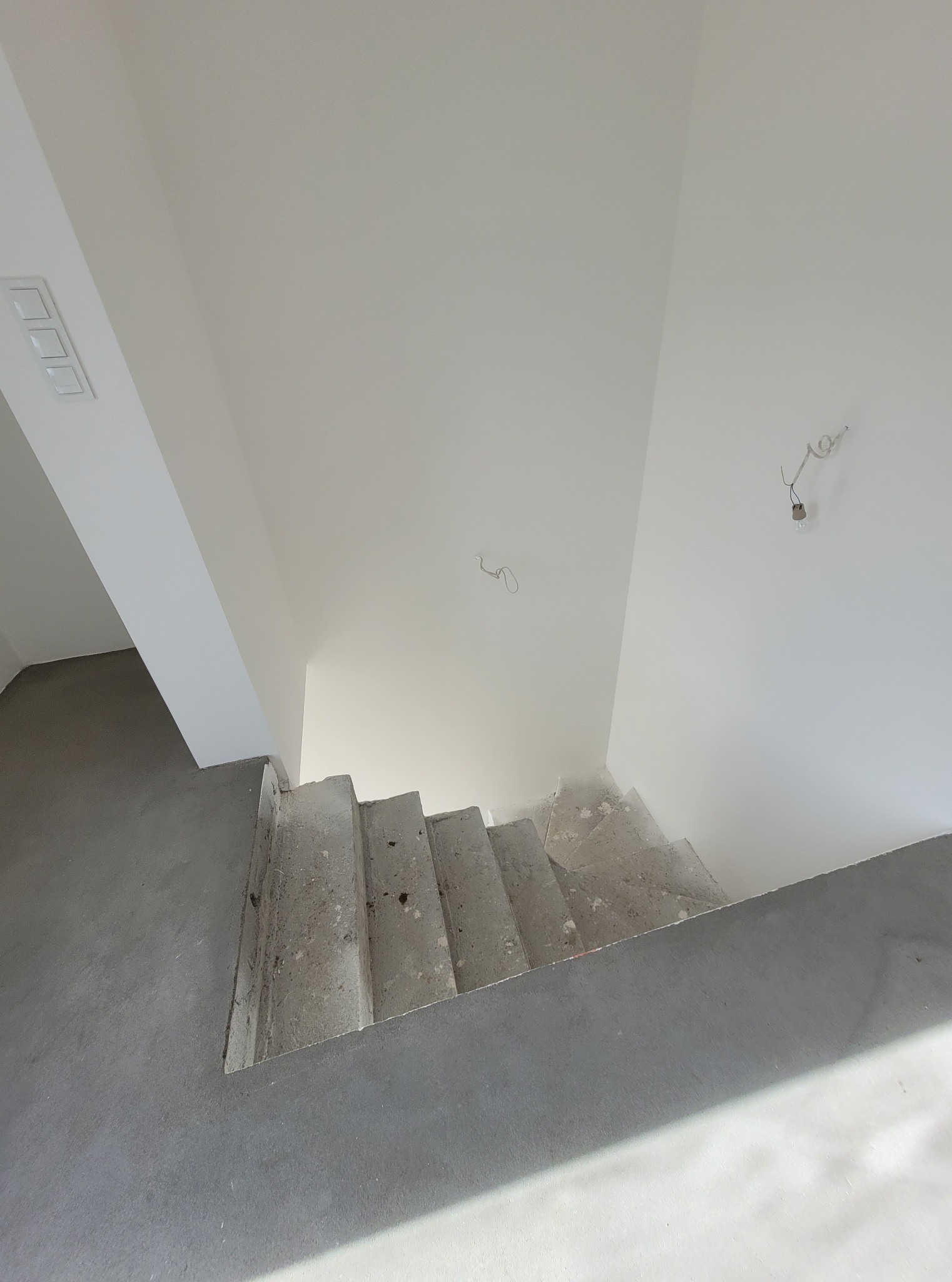 Surowe betonowe schody w jasnym wnętrzu, widok z góry. Świeżo pomalowane ściany na biało, widoczne kable elektryczne. Minimalistyczny design, surowy stan wykończenia.