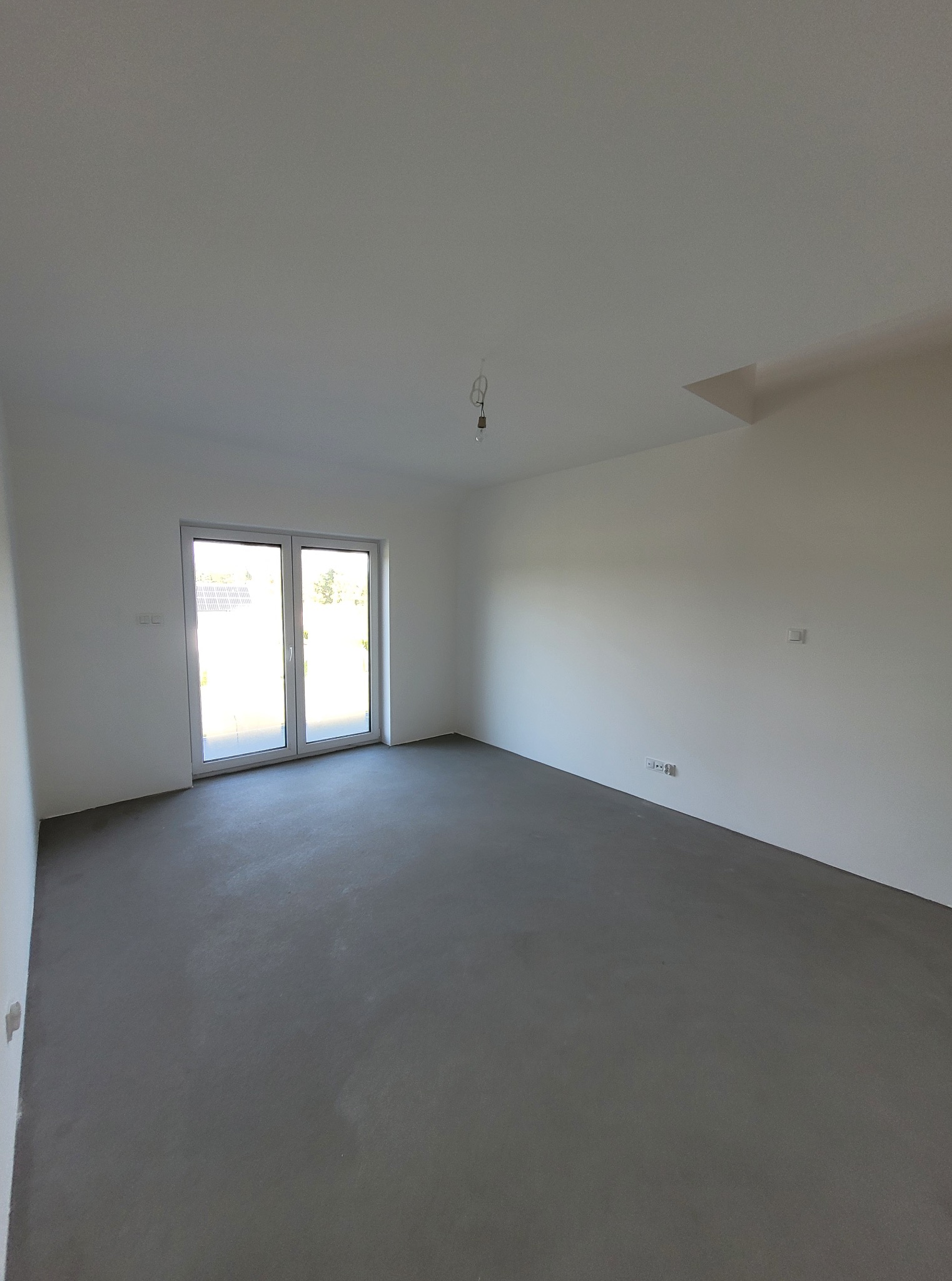 Pusty, świeżo wykończony pokój z szarą wylewką, białymi ścianami i dużym oknem balkonowym. Widoczne gniazdka elektryczne i surowa żarówka.