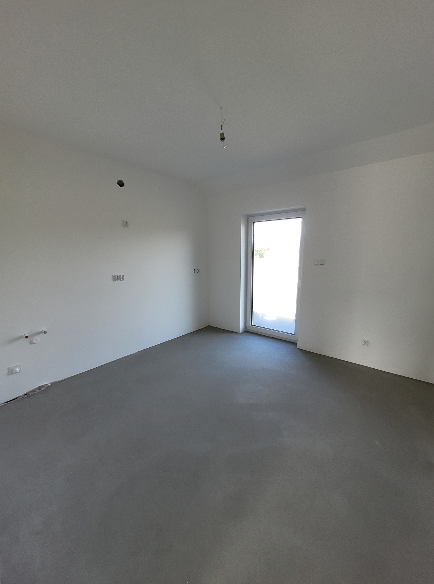 Surowe wnętrze: szare wylewki, białe ściany z instalacjami, okno z widokiem na zieleń. Minimalizm i przygotowanie do dalszych prac wykończeniowych.