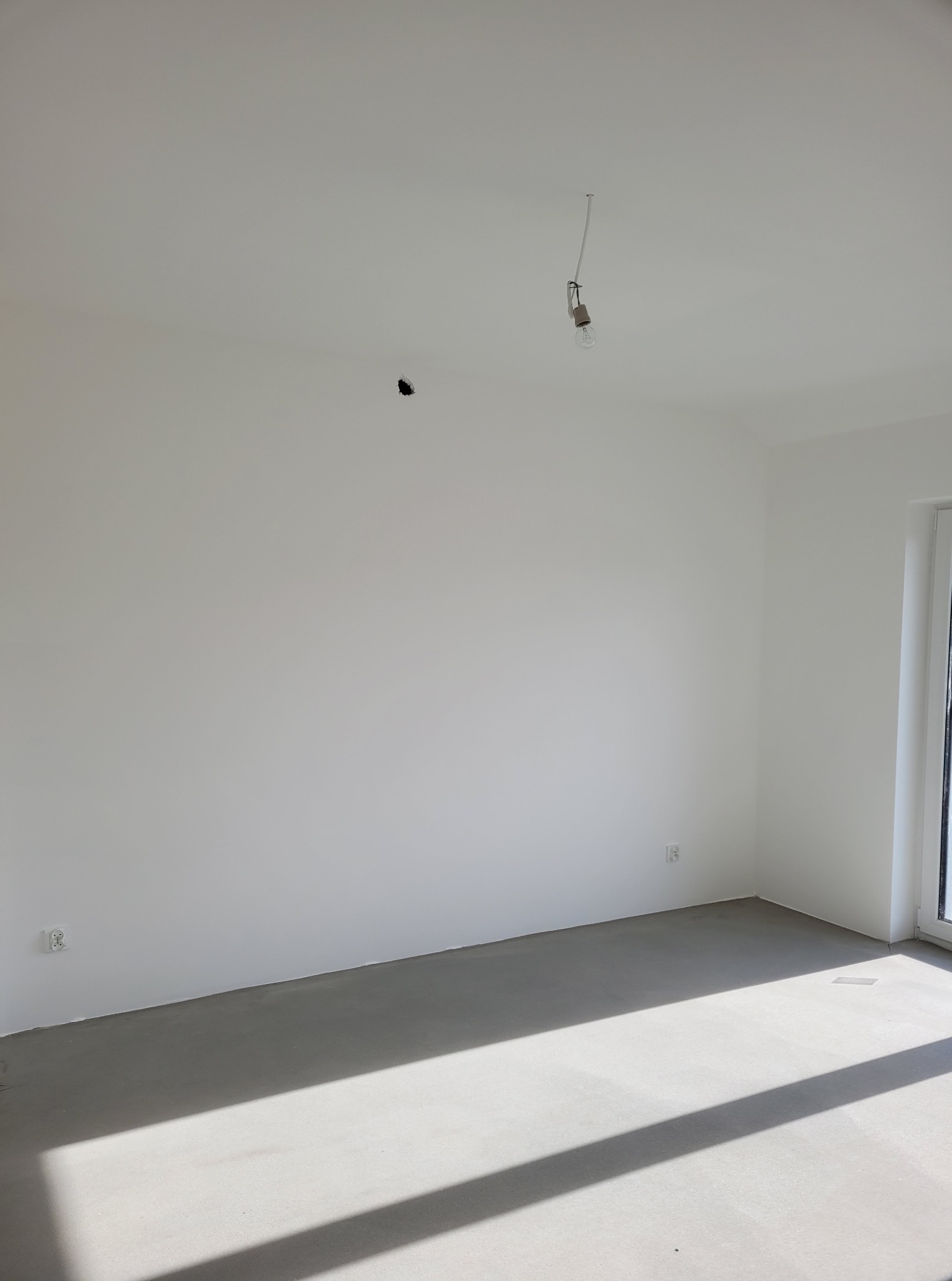 Pusty, świeżo pomalowany pokój z surową podłogą i wiszącą żarówką. Widoczne gniazdka elektryczne i cień padający przez okno. Przygotowanie do wykończenia.