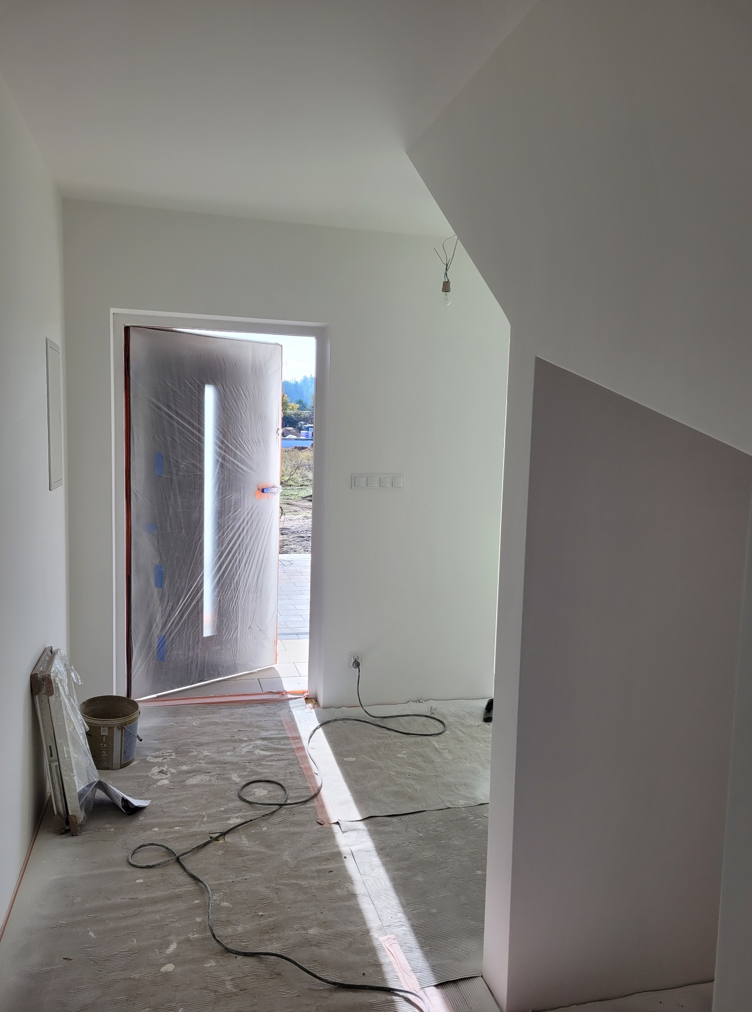 Wnętrze w trakcie prac wykończeniowych: zabezpieczone folią drzwi wejściowe, świeżo pomalowane ściany i podłoga z widocznymi śladami prac. Kabel elektryczny na podłodze.