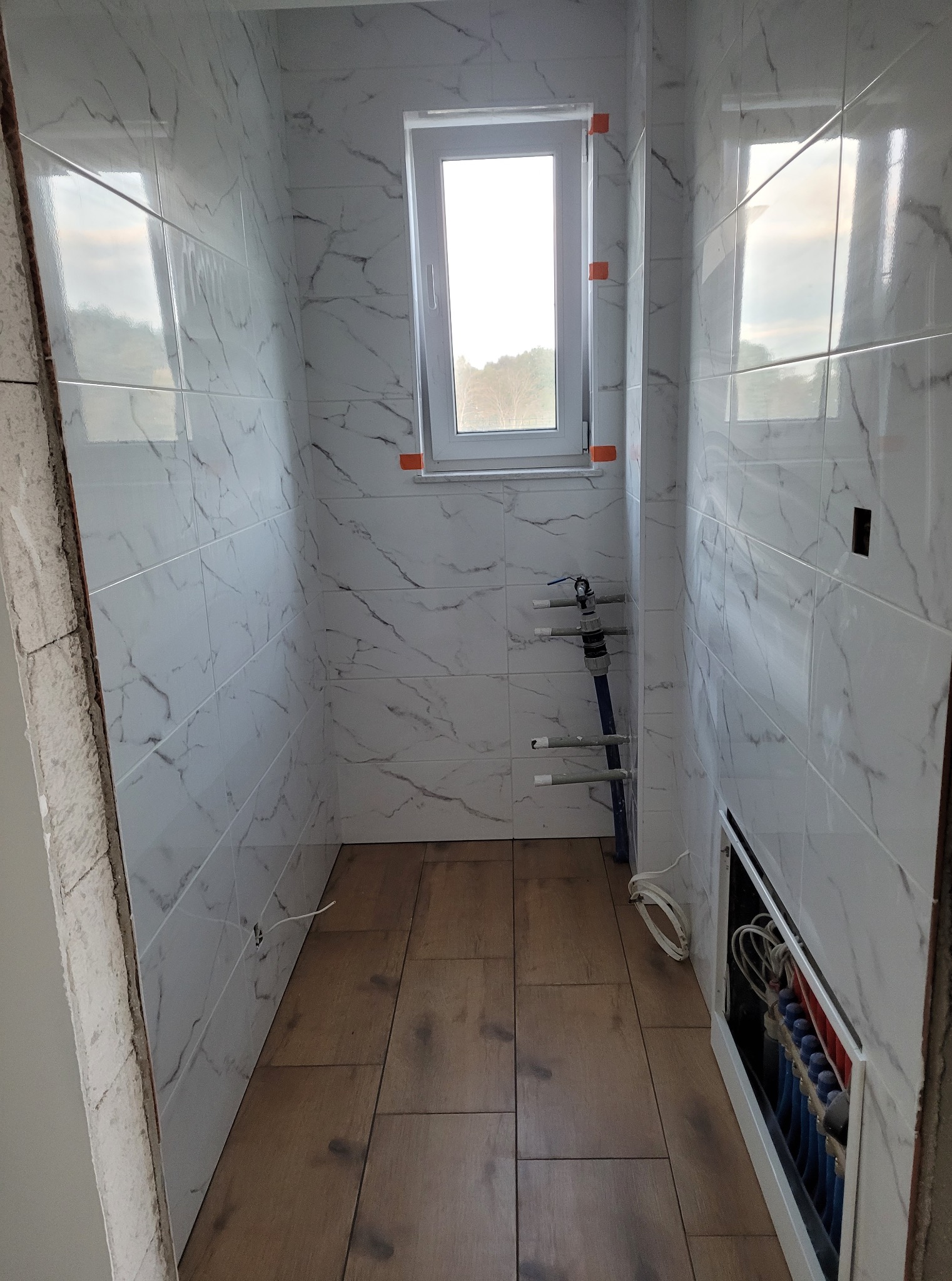 Wnętrze łazienki w Ramleje z białymi płytkami imitującymi marmur i drewnopodobną podłogą. Widoczne instalacje wodne i ogrzewanie podłogowe.