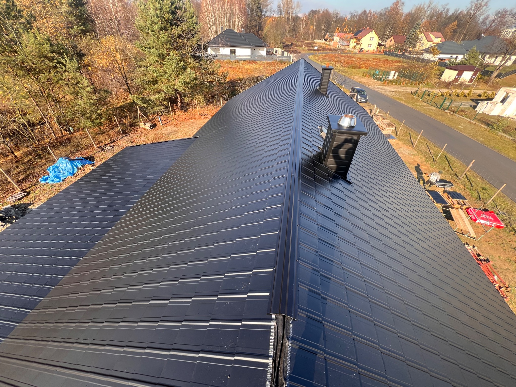 Czarny dach z blachy trapezowej, widok z góry na dom w otoczeniu drzew i zabudowań. Nowoczesne pokrycie dachowe z kominem.
