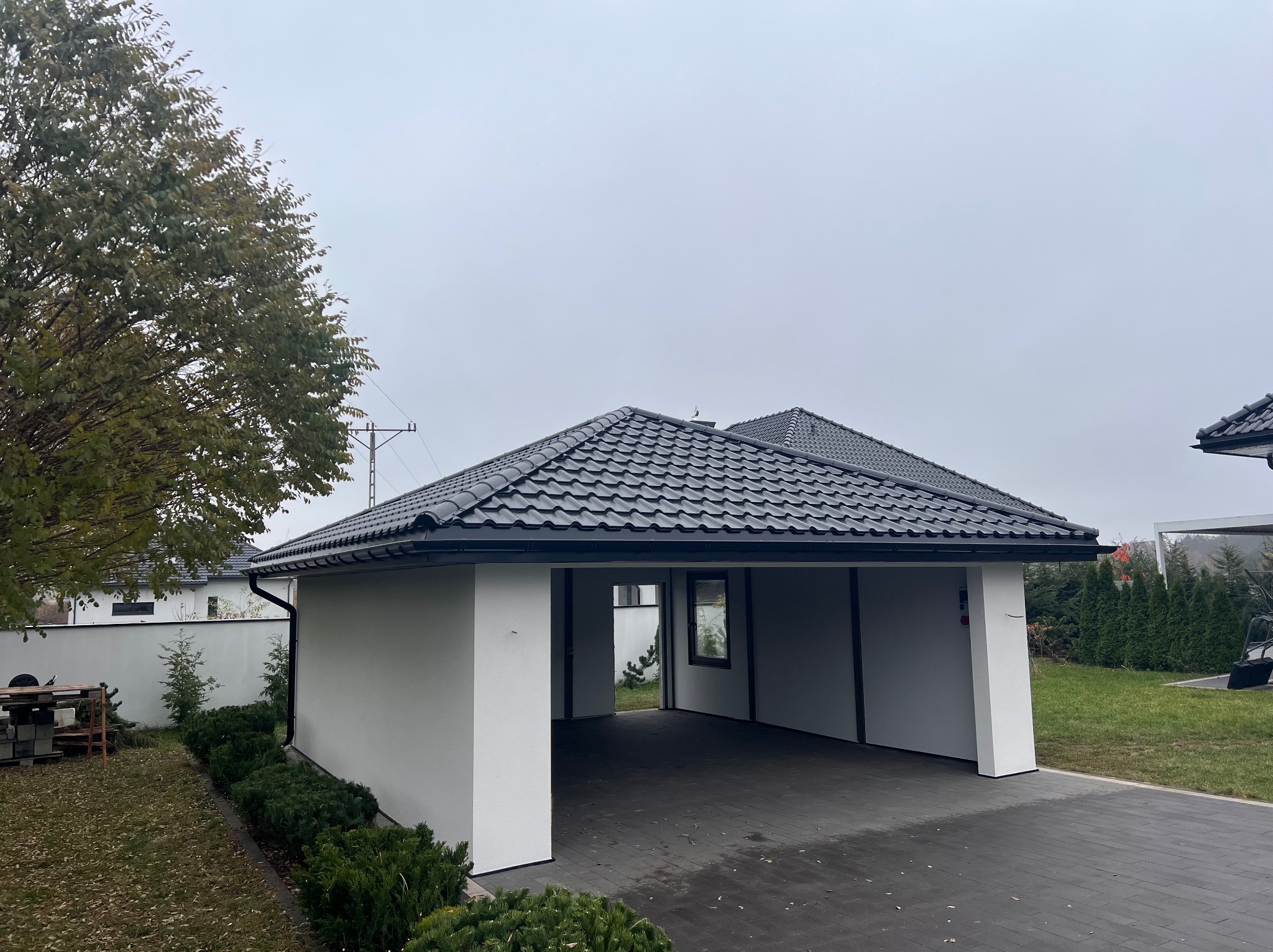 Nowoczesna wiata garażowa z czarną dachówką karpiówką i białymi ścianami, widok z zewnątrz na tle zieleni. Minimalistyczny design, kostka brukowa na podjeździe.