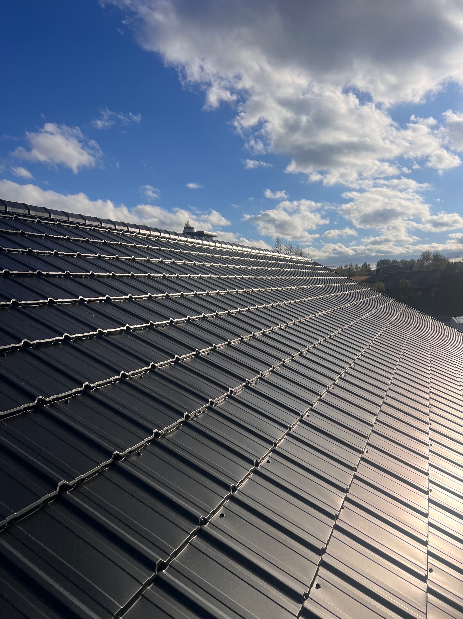 Czarny dach z blachy na tle błękitnego nieba z chmurami. Widoczna faktura blachy i mocowania. Ujęcie z góry, podkreślające regularność wzoru.