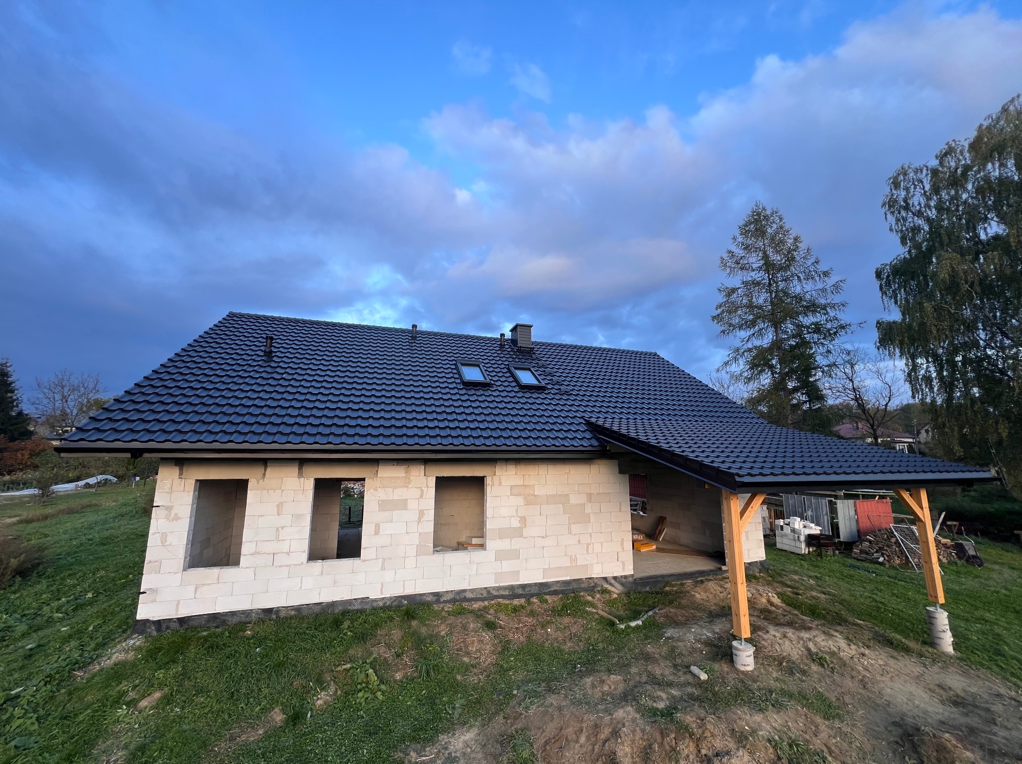Dom w trakcie budowy z czarnym dachem i drewnianą konstrukcją zadaszenia. Widoczne okna dachowe i komin. Budynek z białego bloczka na tle pochmurnego nieba.