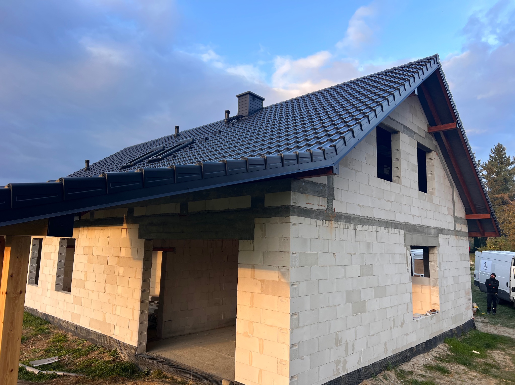 Dom w trakcie budowy z czarnym dachem z dachówki ceramicznej. Widoczne niezadaszone okna i fragment sylwetki osoby w tle. Niebo z chmurami.