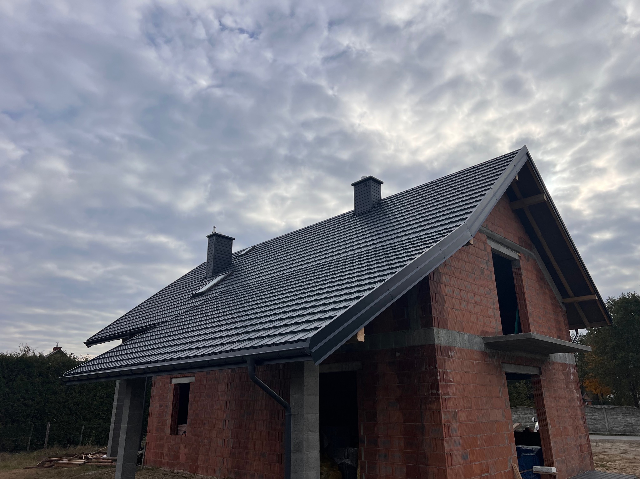 Dom w trakcie budowy z czarnym dachem z dachówki ceramicznej i szarymi rynnami, widok z dołu na tle pochmurnego nieba. Czerwona cegła.