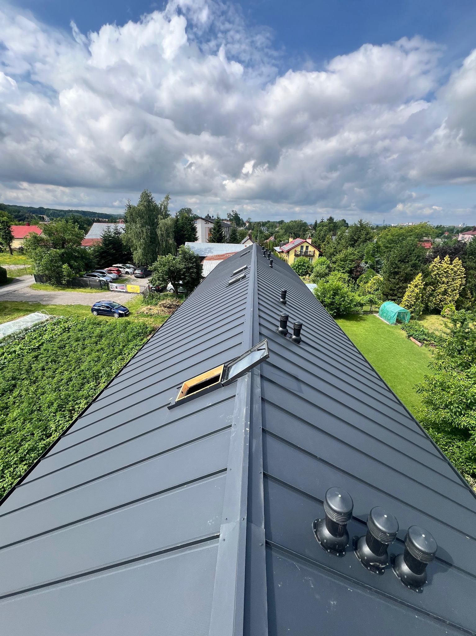 Szary dach z blachy na domu jednorodzinnym, widok z góry, z otwartym oknem dachowym i kominkami wentylacyjnymi. W tle zabudowa jednorodzinna i zieleń.