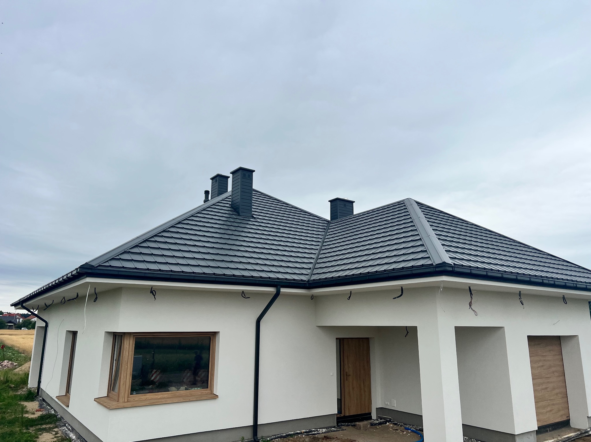 Nowoczesny dom jednorodzinny z ciemnoszarą dachówką ceramiczną i białymi ścianami. Widoczne rynny w kolorze dachu i trzy kominy. Ujęcie z dołu, pochmurne niebo.