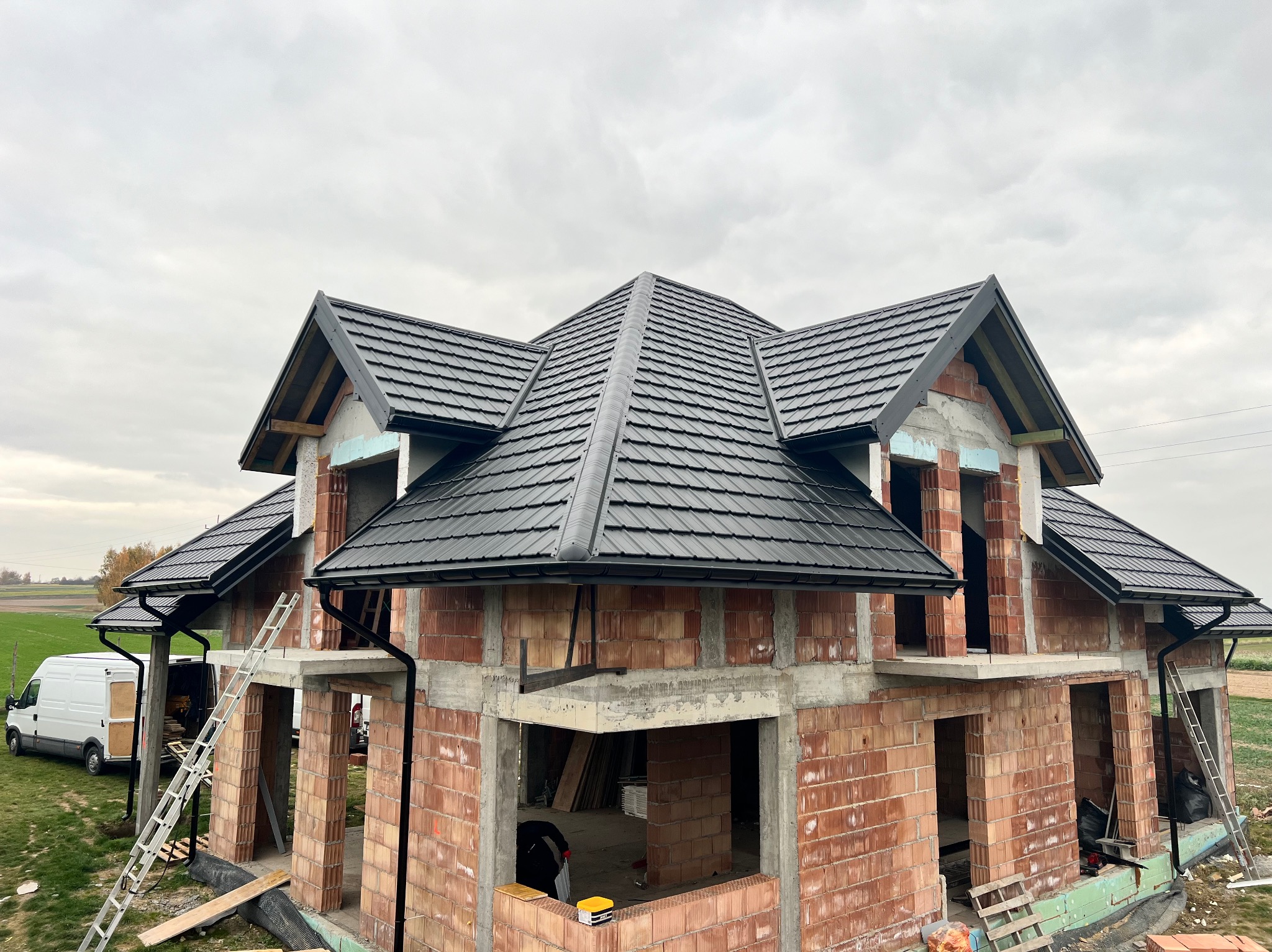Dom w trakcie budowy z czarnym dachem z dachówki ceramicznej. Widoczne rusztowania, drabiny i częściowo wykończona elewacja. Dachówka ułożona w karpiówkę.