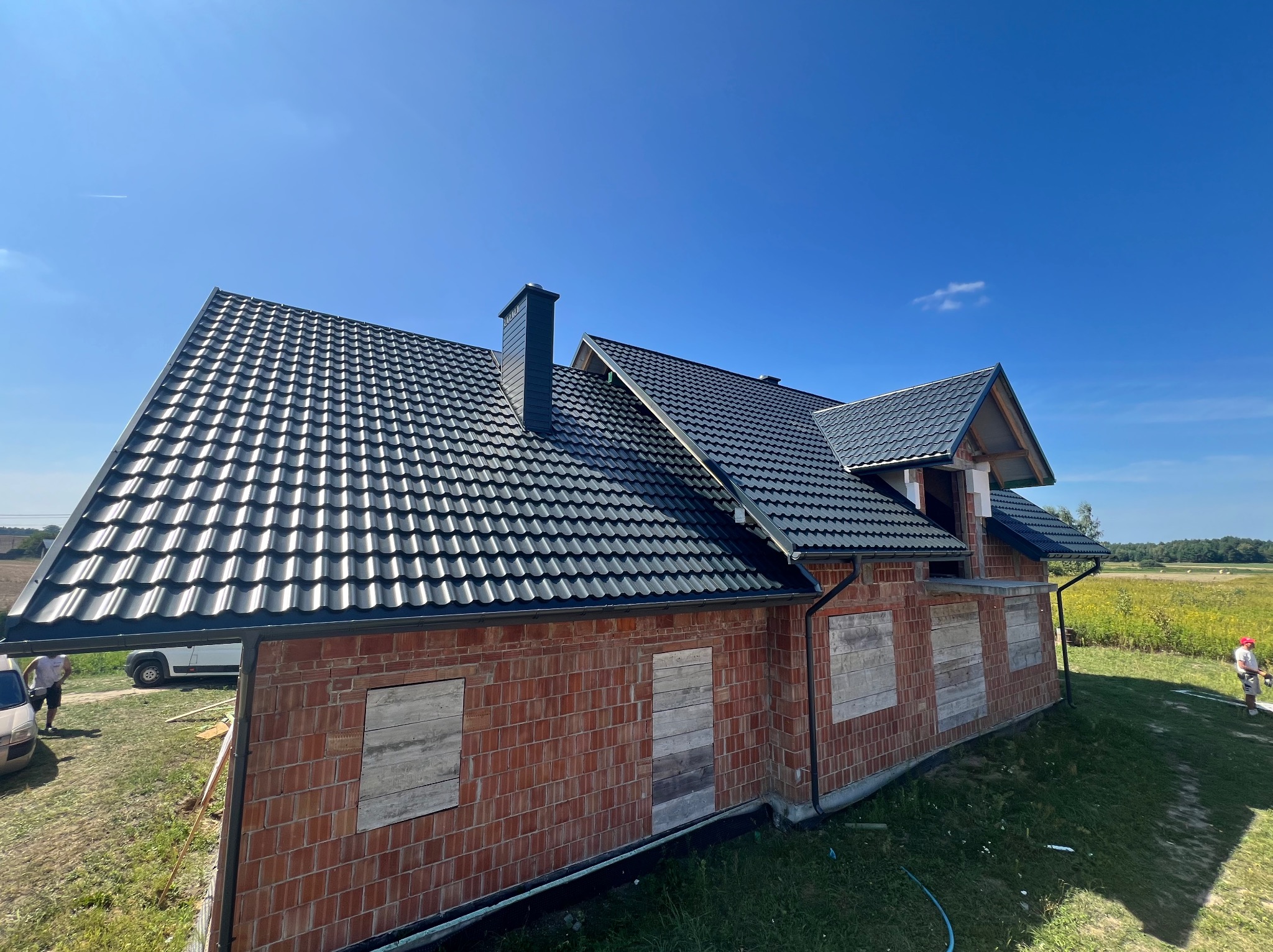 Dom w trakcie budowy z ciemną dachówką i ceglaną elewacją, okna zabite deskami, błękitne niebo w tle. Widok z dołu, podkreślający geometrię dachu.