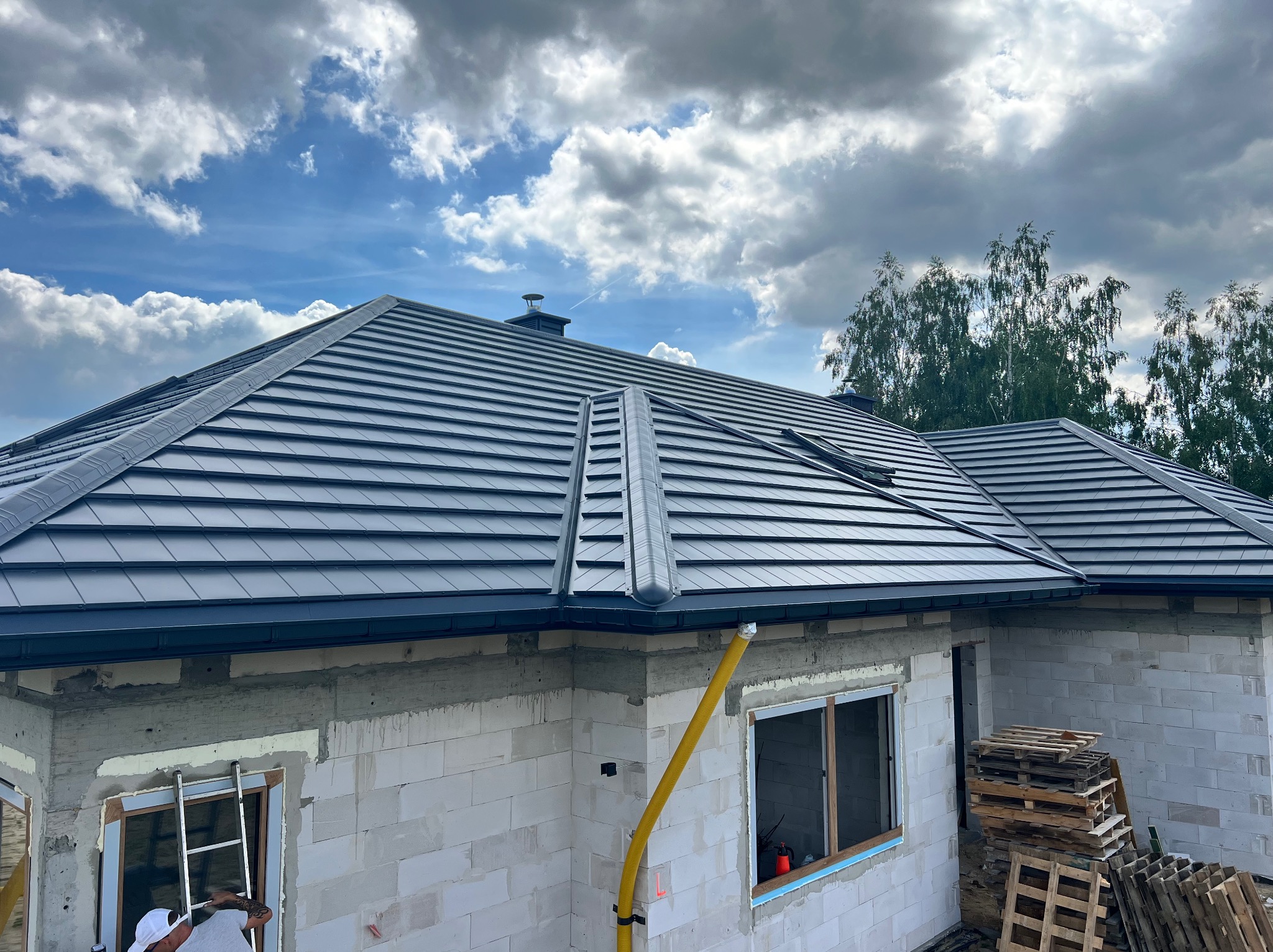 Nowoczesny, grafitowy dach z blachodachówki modułowej na surowym budynku w Górnie, z widoczną rynną i fragmentem drabiny. Błękitne niebo z chmurami w tle.
