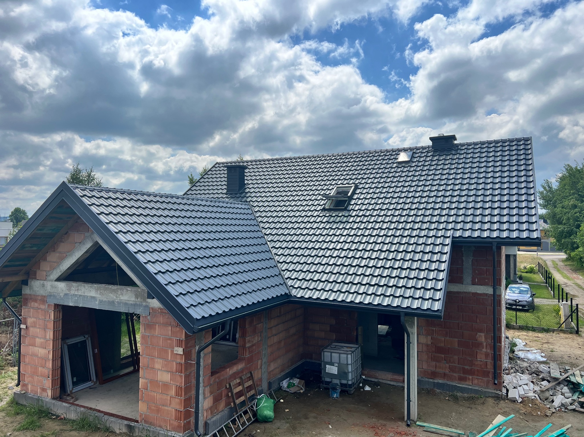 Dom w trakcie budowy z nowym, grafitowym dachem z dachówki modułowej i oknem dachowym, na tle błękitnego nieba z chmurami. Widoczne obróbki blacharskie.