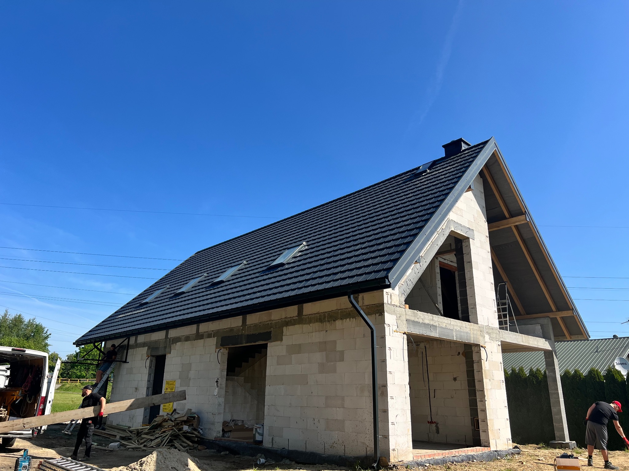 Dom w trakcie budowy z czarnym dachem i oknami dachowymi. Pracownicy budowlani w trakcie pracy. Błękitne niebo w tle. Widoczne rynny.