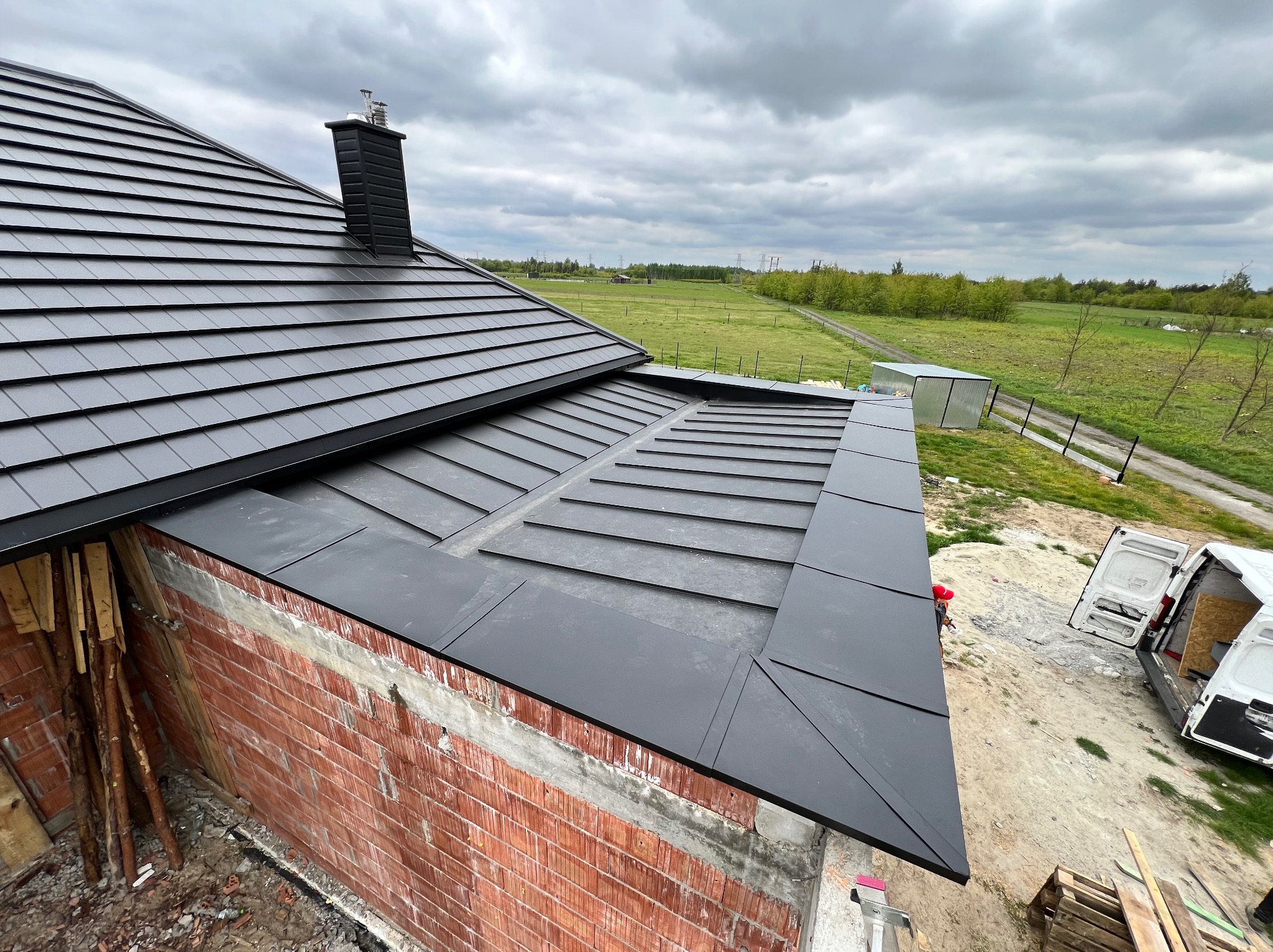 Czarny dach z blachy na budynku w trakcie budowy, widok z góry, z kominem i fragmentem ceglanej ściany. W tle zielone pola i pochmurne niebo.