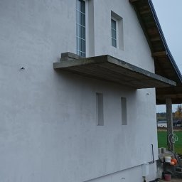 Usługi ogólnobudowlane Łukasz Sosnowski - Boczny widok domu z jasną elewacją, widoczne okna, betonowy balkon i fragment dachu. Teren wokół domu częściowo zagospodarowany, w tle zieleń.