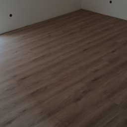 Usługi ogólnobudowlane Łukasz Sosnowski - Ukończona podłoga z paneli w odcieniach drewna, widoczna w jasnym pomieszczeniu z białymi ścianami i oknem z roletą. Świeżo wykończone ściany.