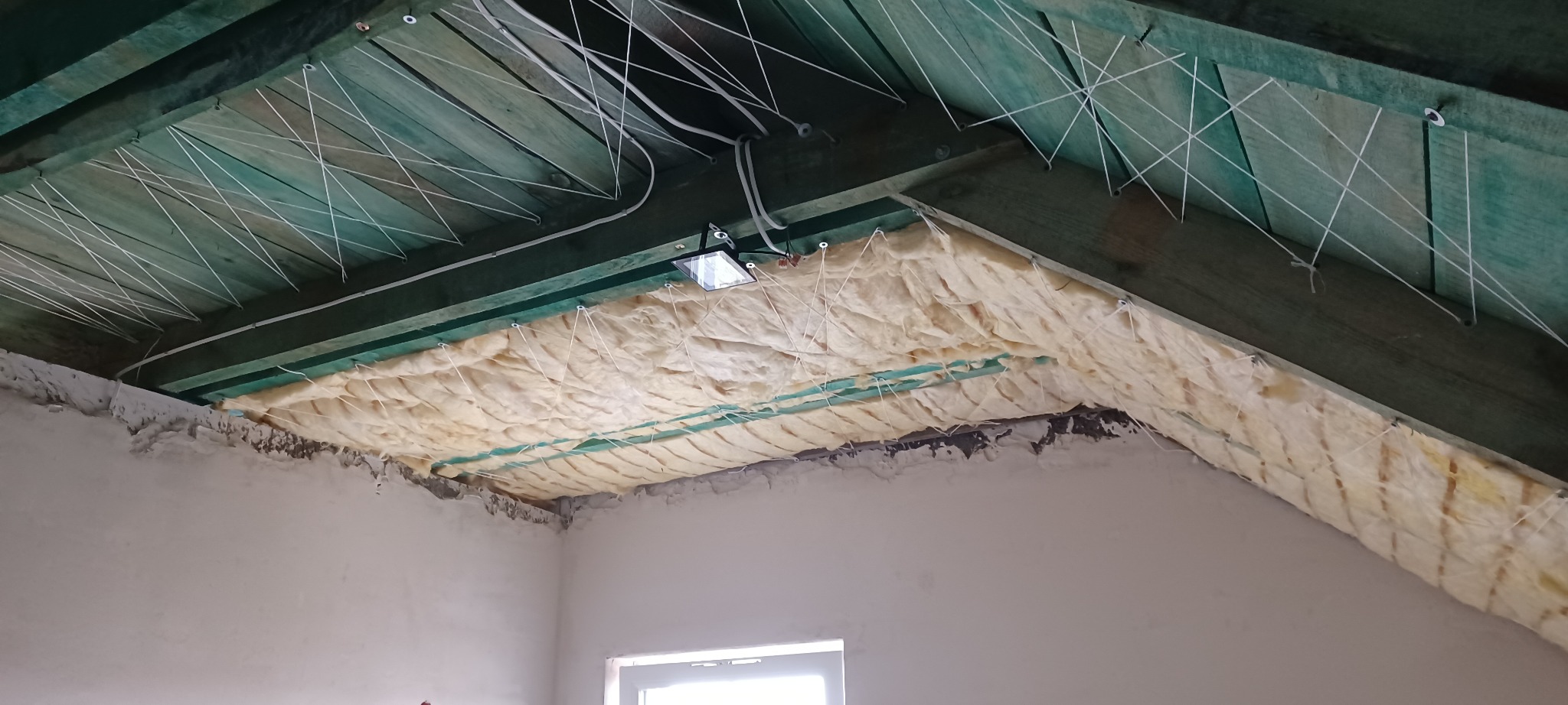 Poddasze z widoczną izolacją z wełny mineralnej, zabezpieczoną sznurkami przymocowanymi do drewnianej konstrukcji dachu. Widoczne oświetlenie robocze.