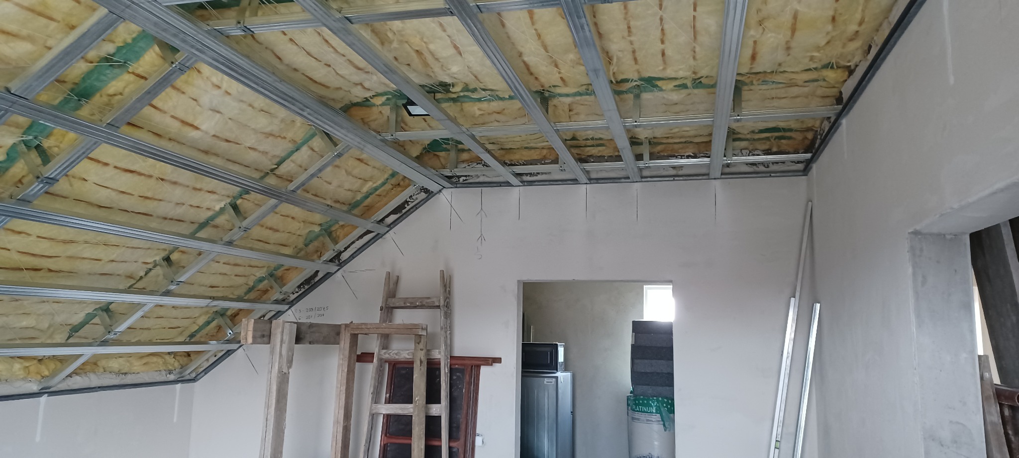 Skośny sufit podwieszany z izolacją z wełny mineralnej, widoczne metalowe profile i fragment ściany z zaznaczonymi liniami. W tle widoczne drzwi i sprzęt AGD.